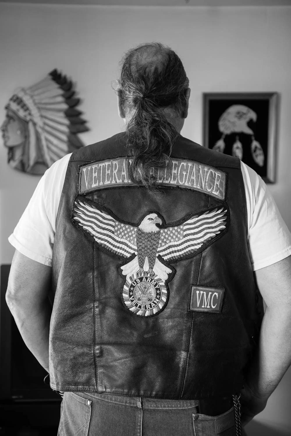 Wearing his motorcycle vest, Michael stands before two wood carvings that his father had made. Michael's father has passed away. 