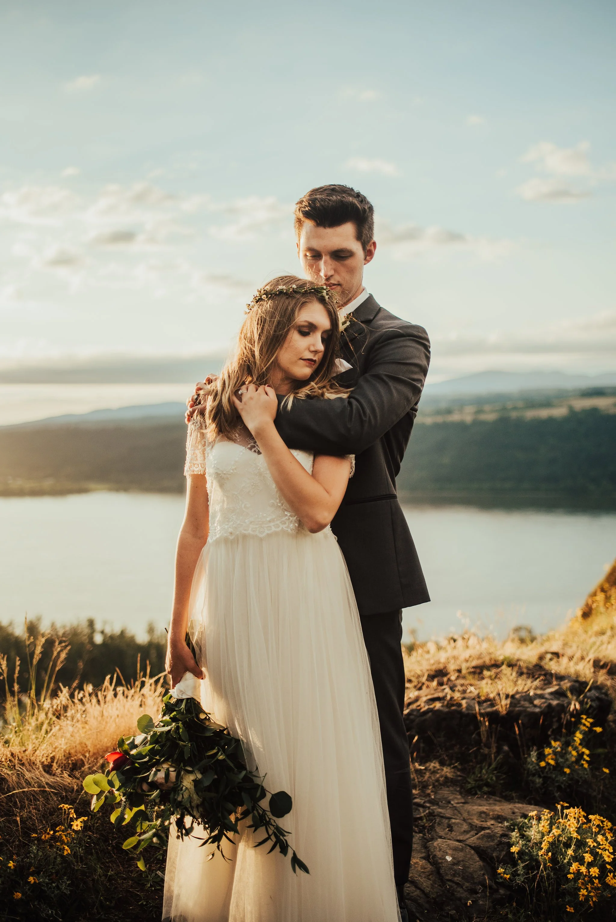 A newlywed couple stands close together outdoors during sunset in the Columbia Gorge outside of Portland, OR. They are on a rocky hill with a scenic lake and mountains in the background.