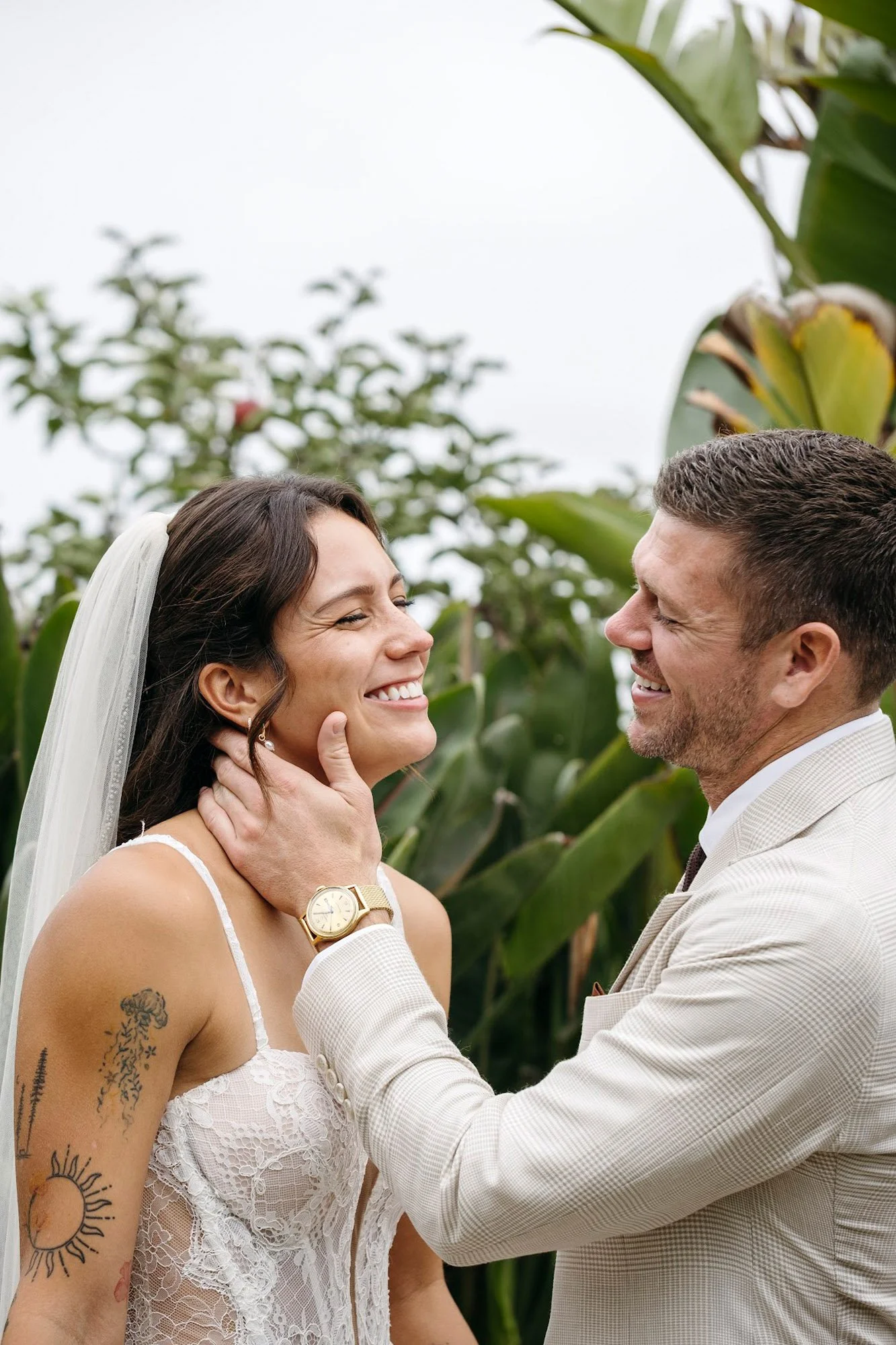 A man and woman smiling during a wedding photo, with the man holding the woman's face, in a lush outdoor setting with green plants.