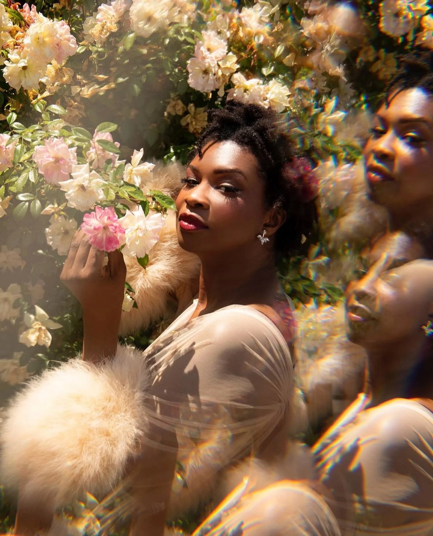 A woman in a beige dress and fur sleeves poses among blooming pink and white flowers, with a mirrored effect creating a duplicate of her face on the right side.