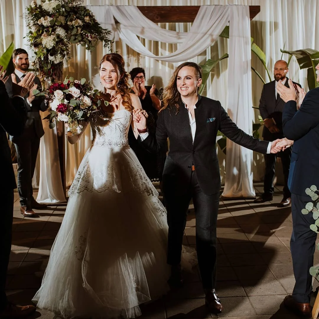 A bride in a white lace wedding gown holding a bouquet of flowers, smiling and holding hands with another woman in a black suit, at a wedding ceremony with guests clapping and a decorated backdrop with draped white fabric and greenery.