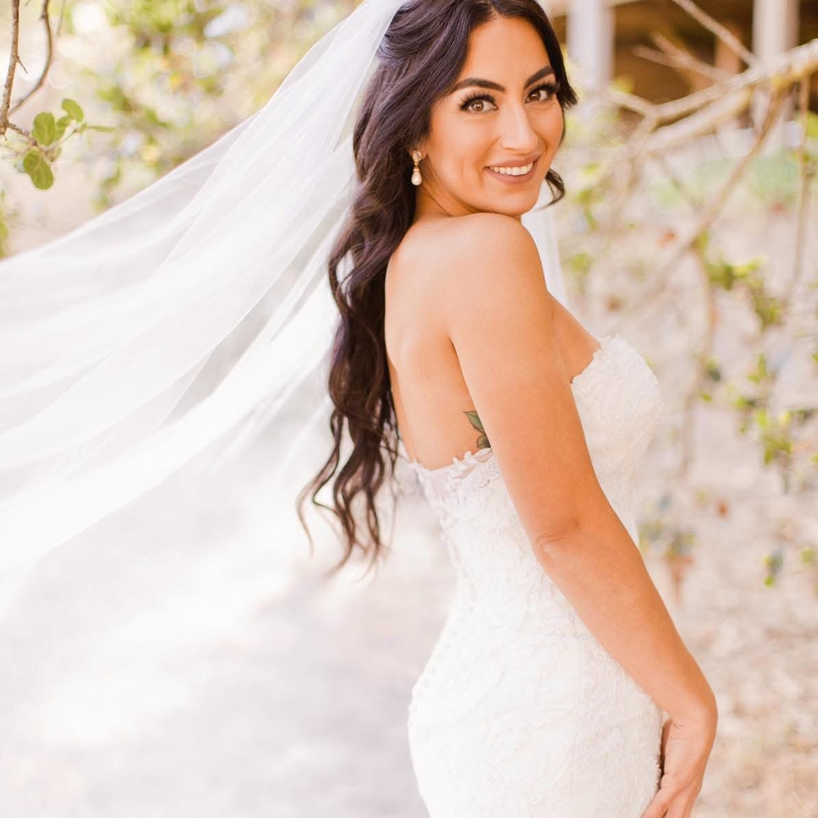 Smiling bride in white wedding dress with veil, outdoor setting with greenery and branches in the background.