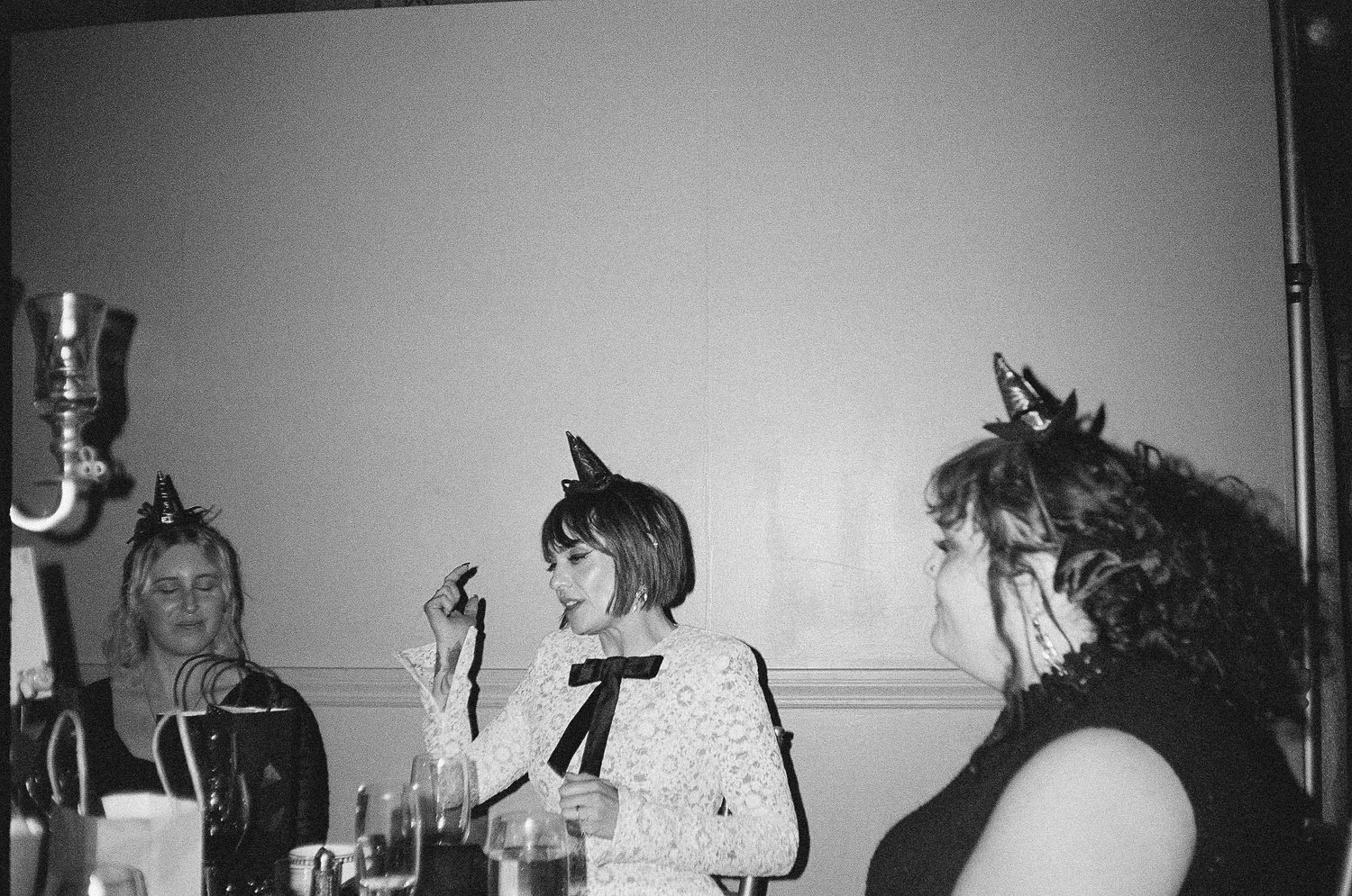 Women celebrating a birthday at a party, wearing cone-shaped party hats and themed accessories, sitting at a table with drinks.