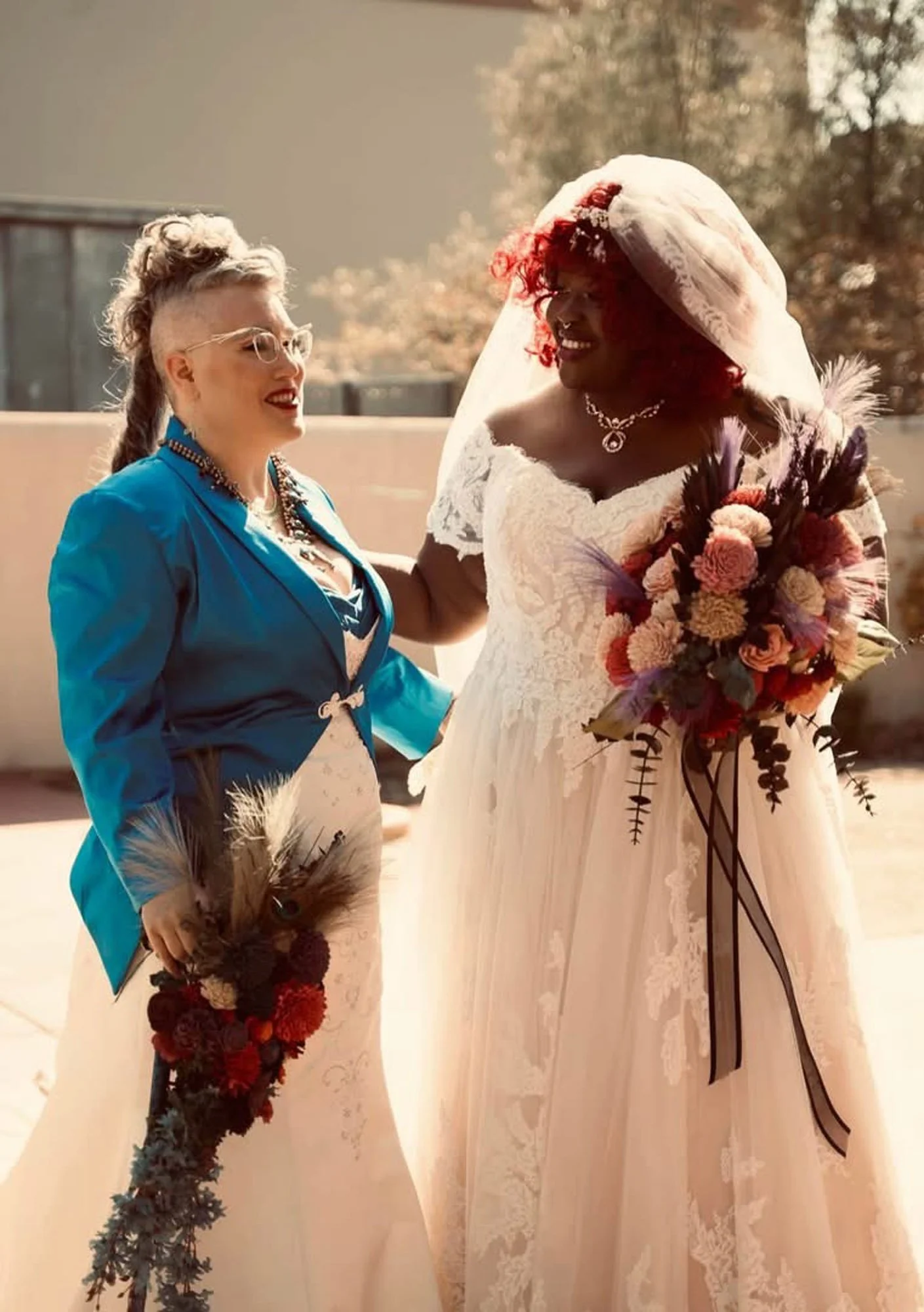 Two women, one in a blue jacket and glasses, the other in a white wedding dress, smiling and holding bouquets, standing outdoors with a tree and building in the background.