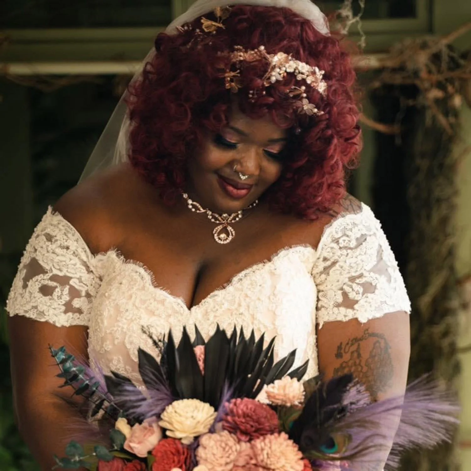 A woman with dark skin, curly red hair, and a sweetheart lace wedding dress is holding a bouquet of flowers and smiling.