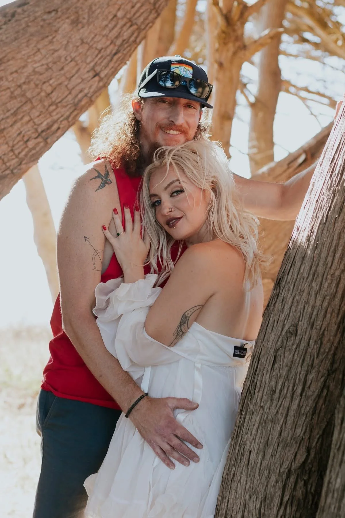 A couple standing outdoors among trees, with the man wearing a sleeveless red shirt, sunglasses on his cap, and the woman in a white off-shoulder dress, both embracing and looking at the camera.