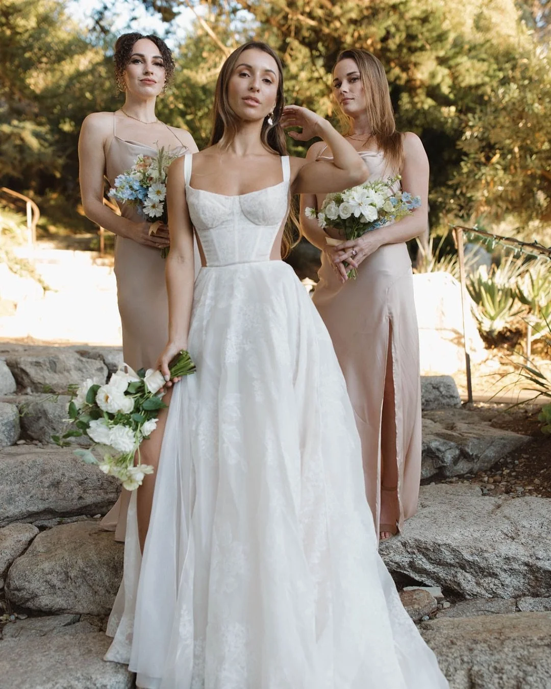 Three women standing outdoors on rocks during a wedding, with the woman in the center wearing a white wedding gown, holding a bouquet, and the women on either side wearing matching beige dresses and holding bouquets.