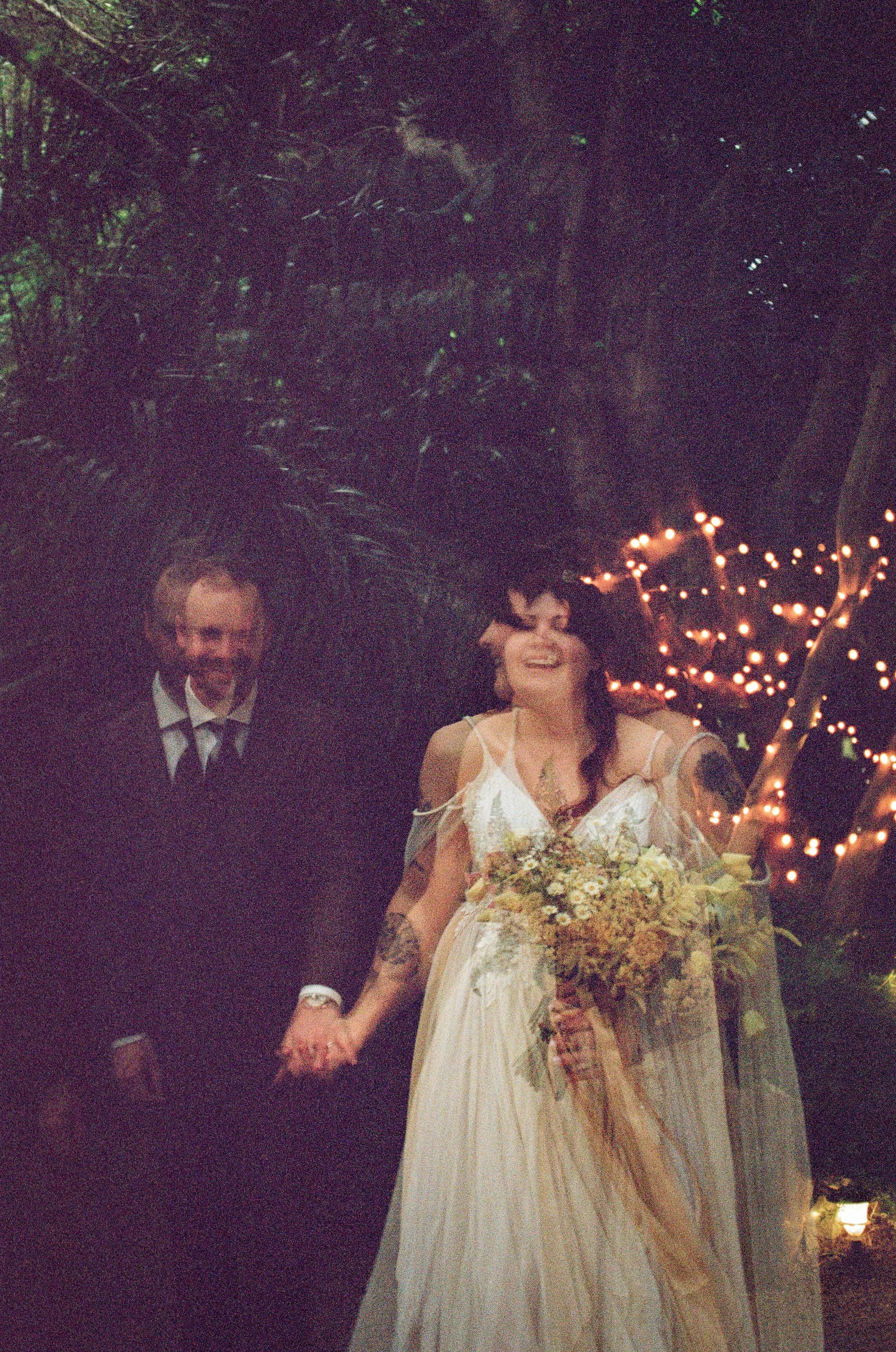 A couple dressed for a wedding, holding hands, standing outdoors at night with string lights in the background.