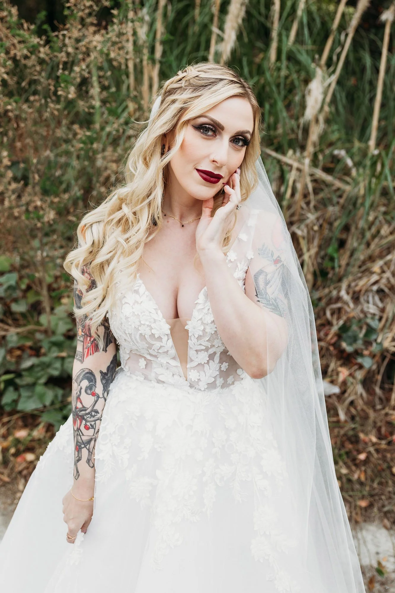 A woman in a white wedding dress with lace floral details, standing outdoors near tall grasses and bushes. She has long, wavy blonde hair, dark makeup, and tattoos on her left arm. She is touching her face with her right hand and looking at the camera.