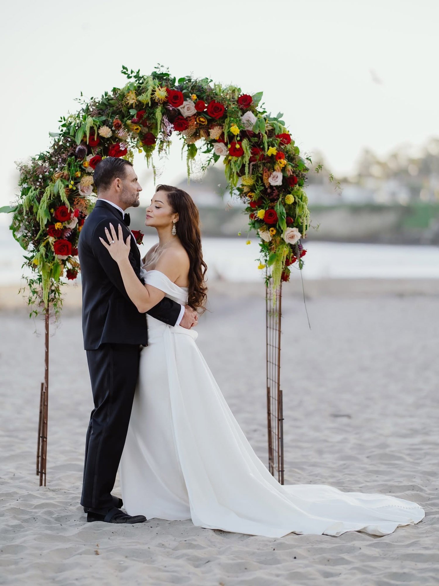 A bride and groom in wedding attire embracing on a sandy beach in front of a colorful flower arch during their wedding ceremony.
