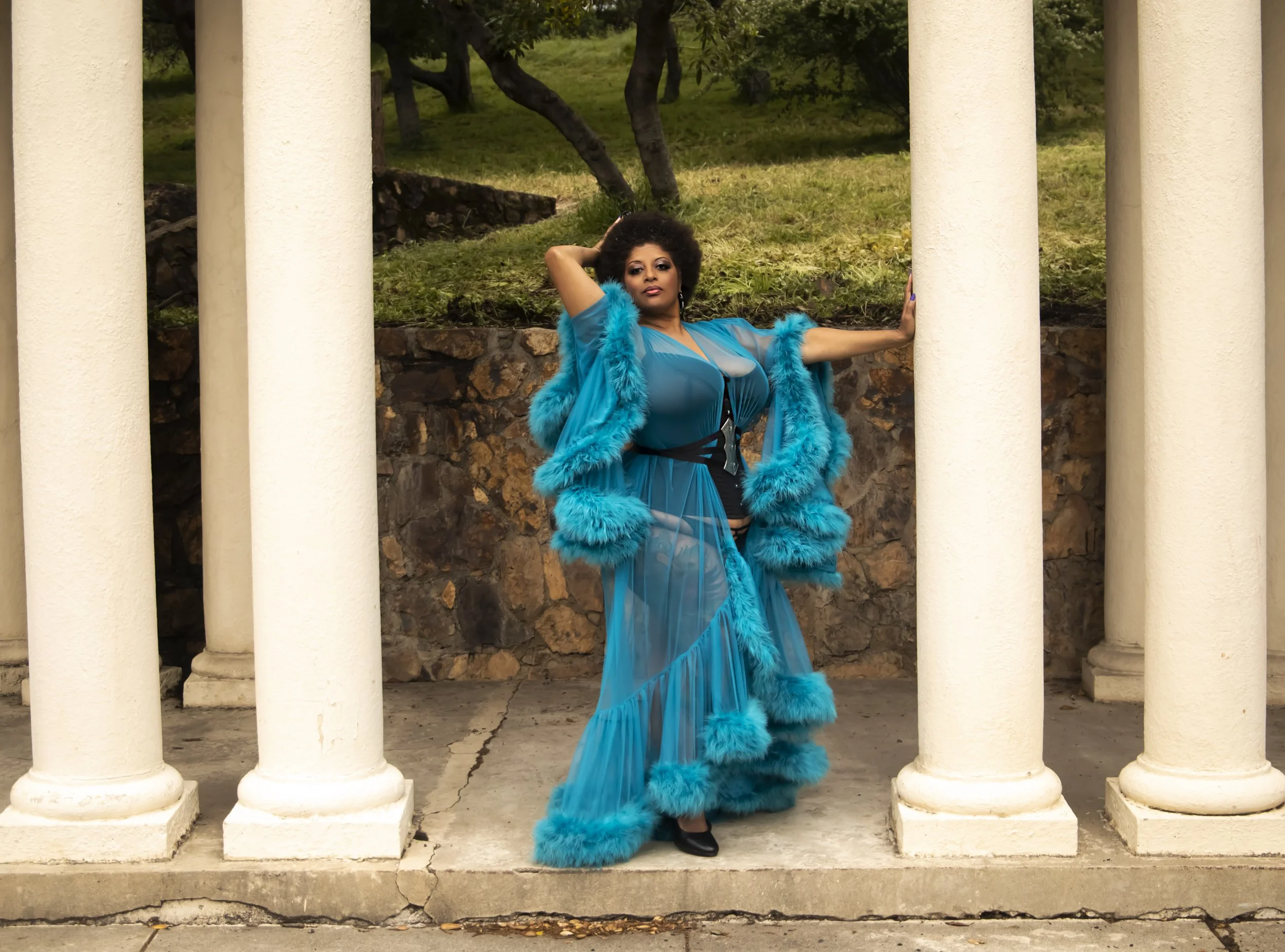 A woman in a sheer blue dress and matching fur coat posing between white columns outdoors.