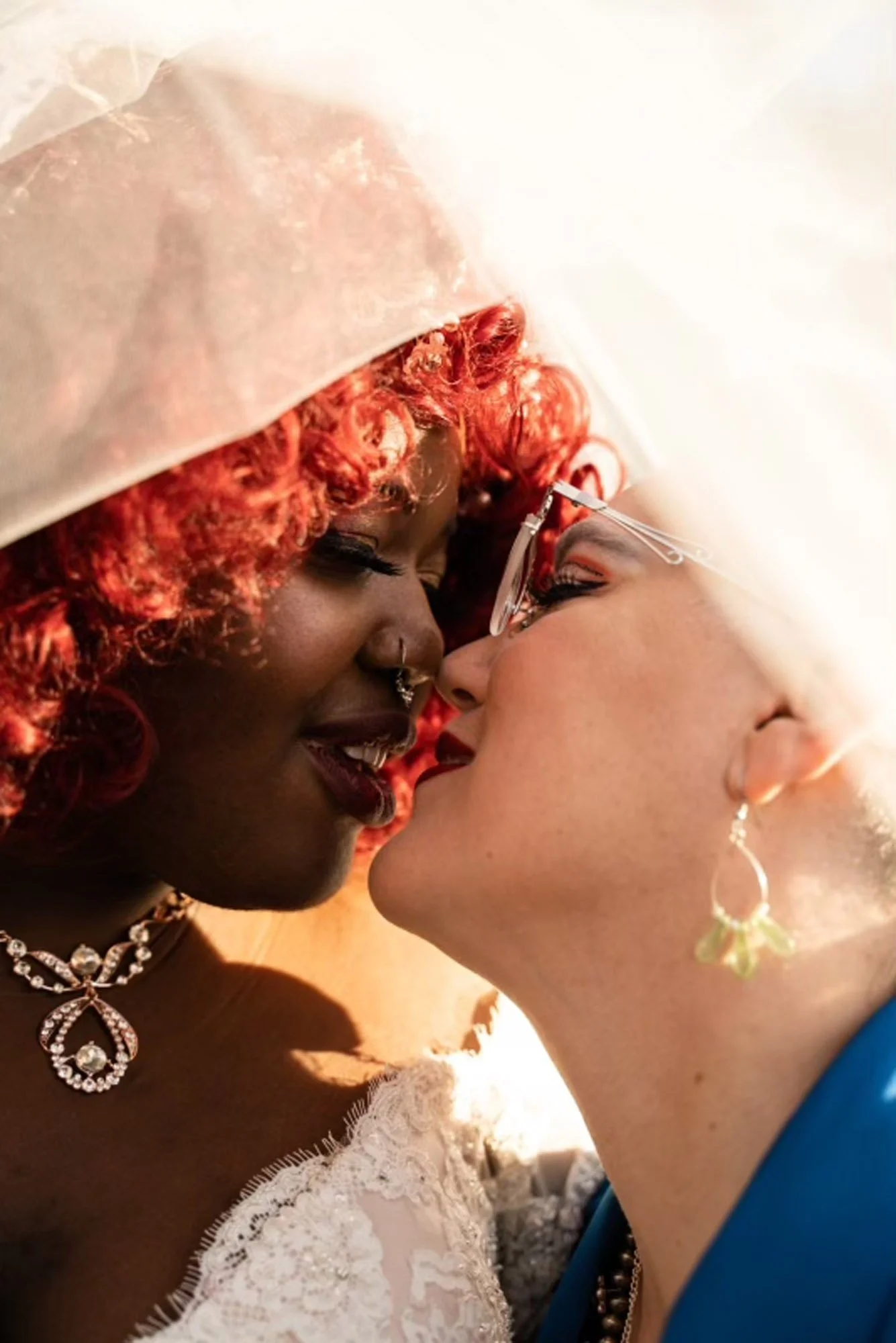 Two women with different skin tones are close together, about to kiss. One has curly red hair, glasses, and a lace dress, while the other has short blonde hair, earrings, and a blue outfit.