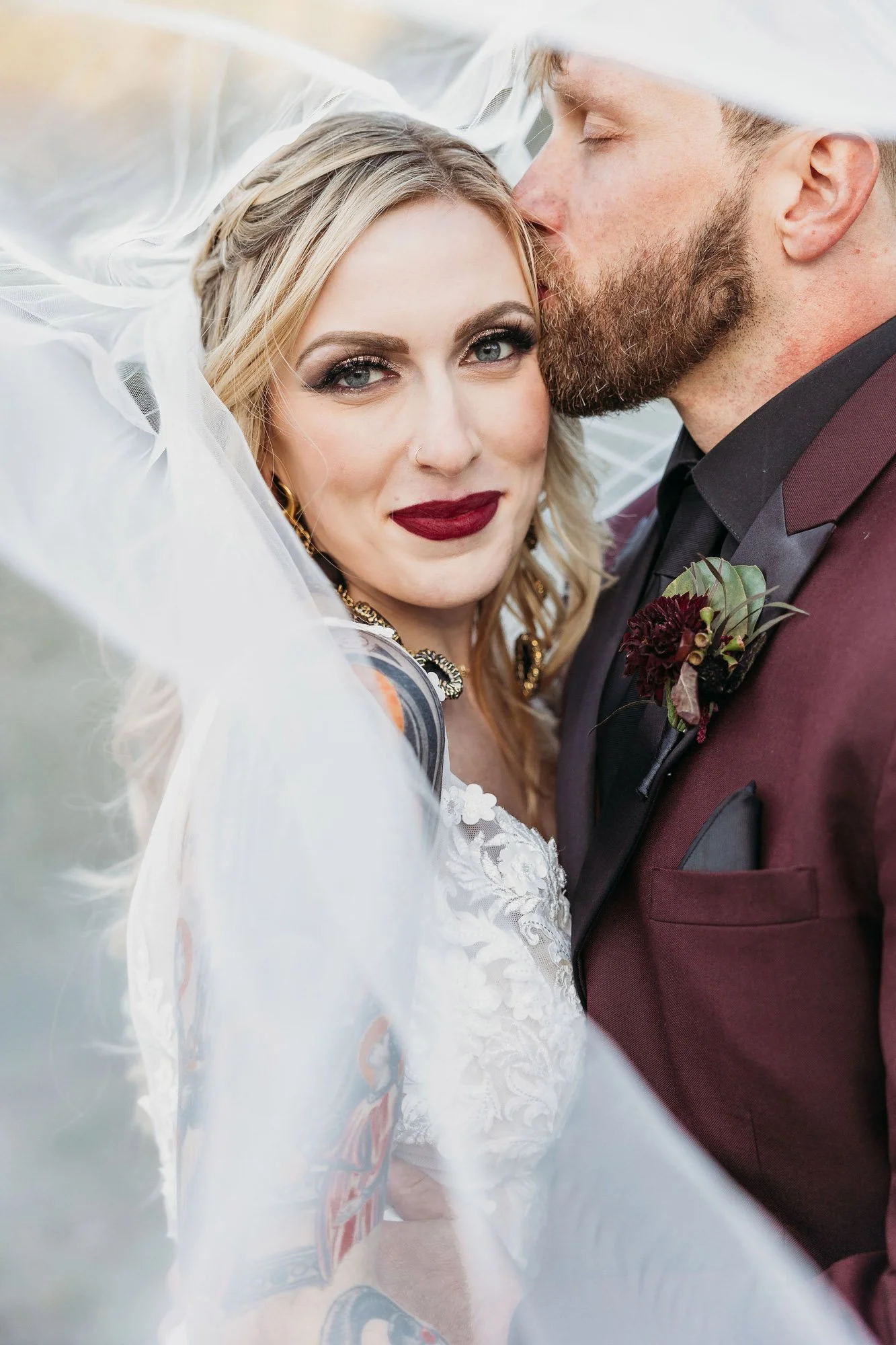 A bride and groom close-up on their wedding day, with the groom kissing the bride's forehead. The bride is wearing a white lace dress with tattoos on her arm and bold makeup. The groom is in a burgundy tuxedo with a boutonniere.