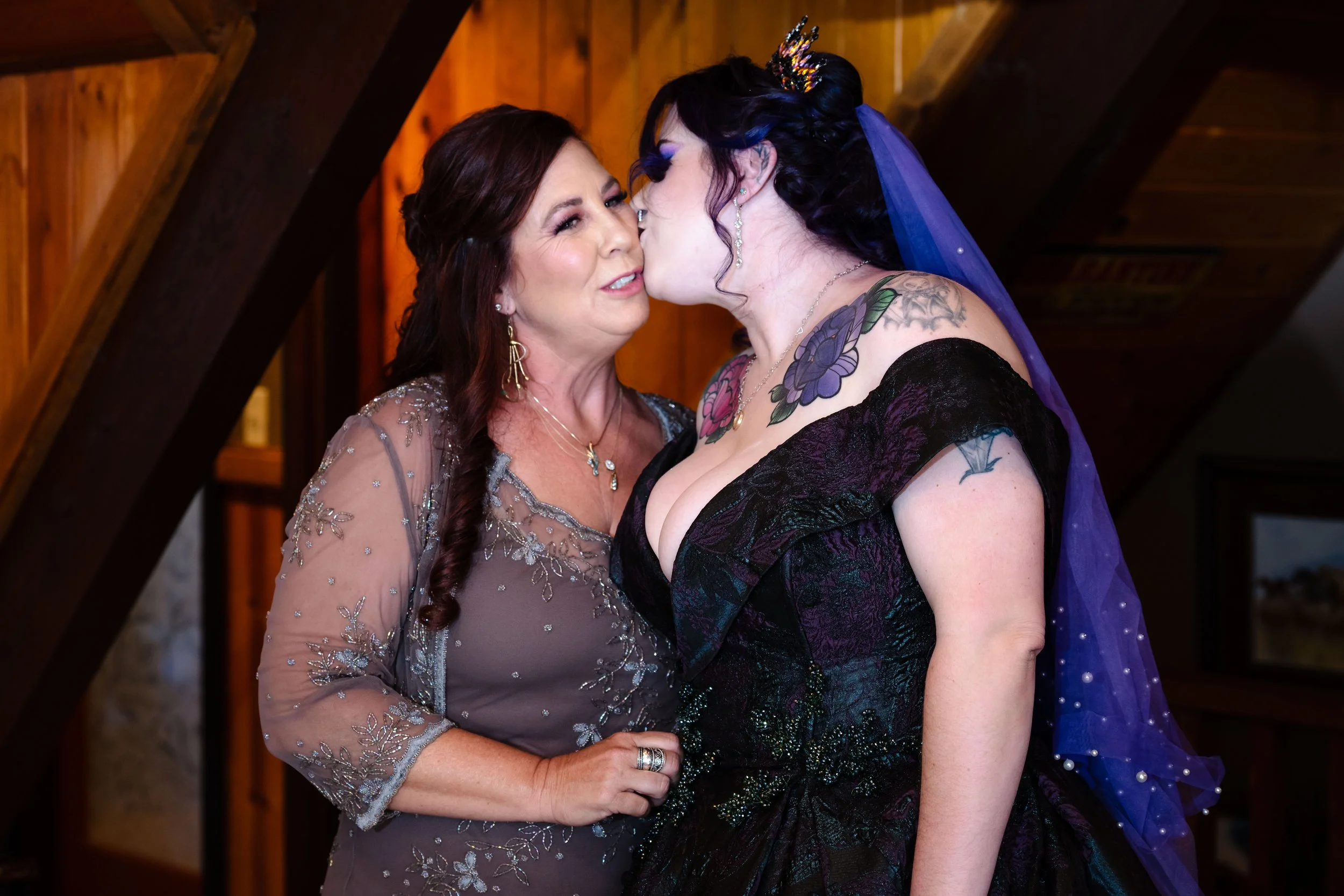 A gothic bride gives the mother of the bride a kiss on the cheek. The mother of the bride has soft glam colorful makeup 