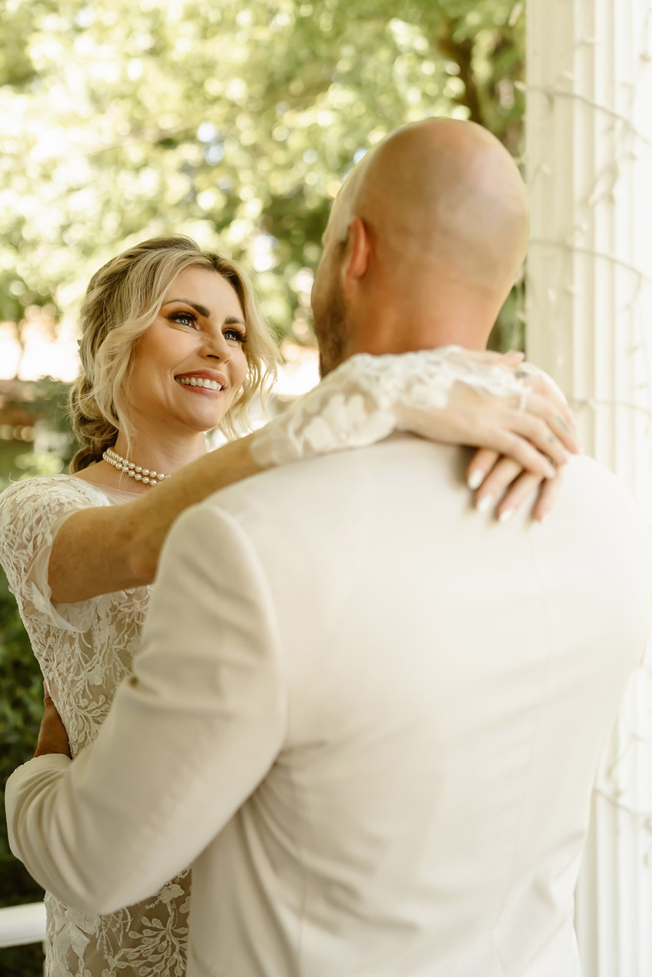 A woman with blonde hair and a pearl necklace smiling at a man with a bald head, both dressed in wedding attire, outside in a green environment.