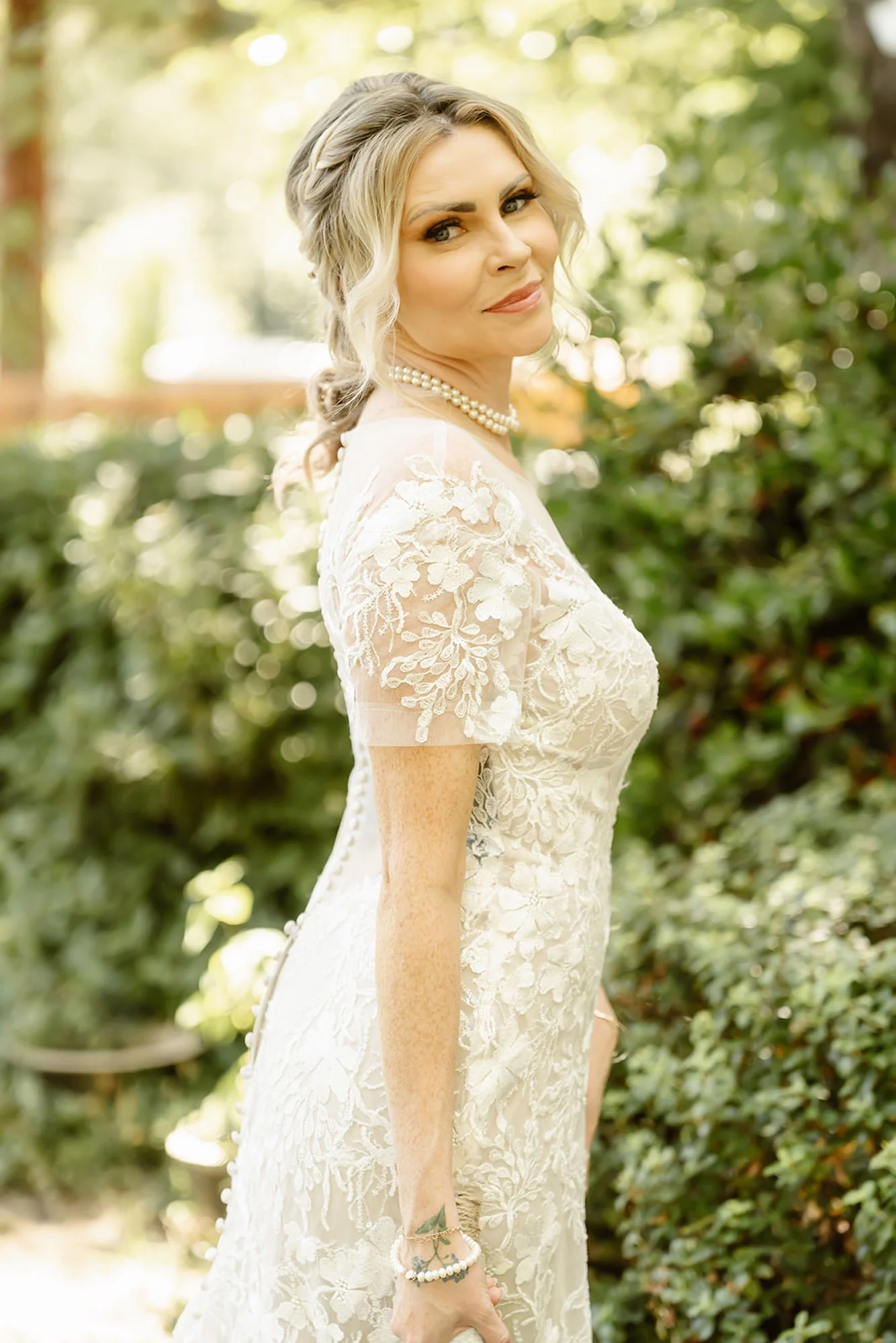 A woman in a lace wedding dress standing outdoors, wearing a pearl necklace and bracelet, with wavy hair and a gentle smile.
