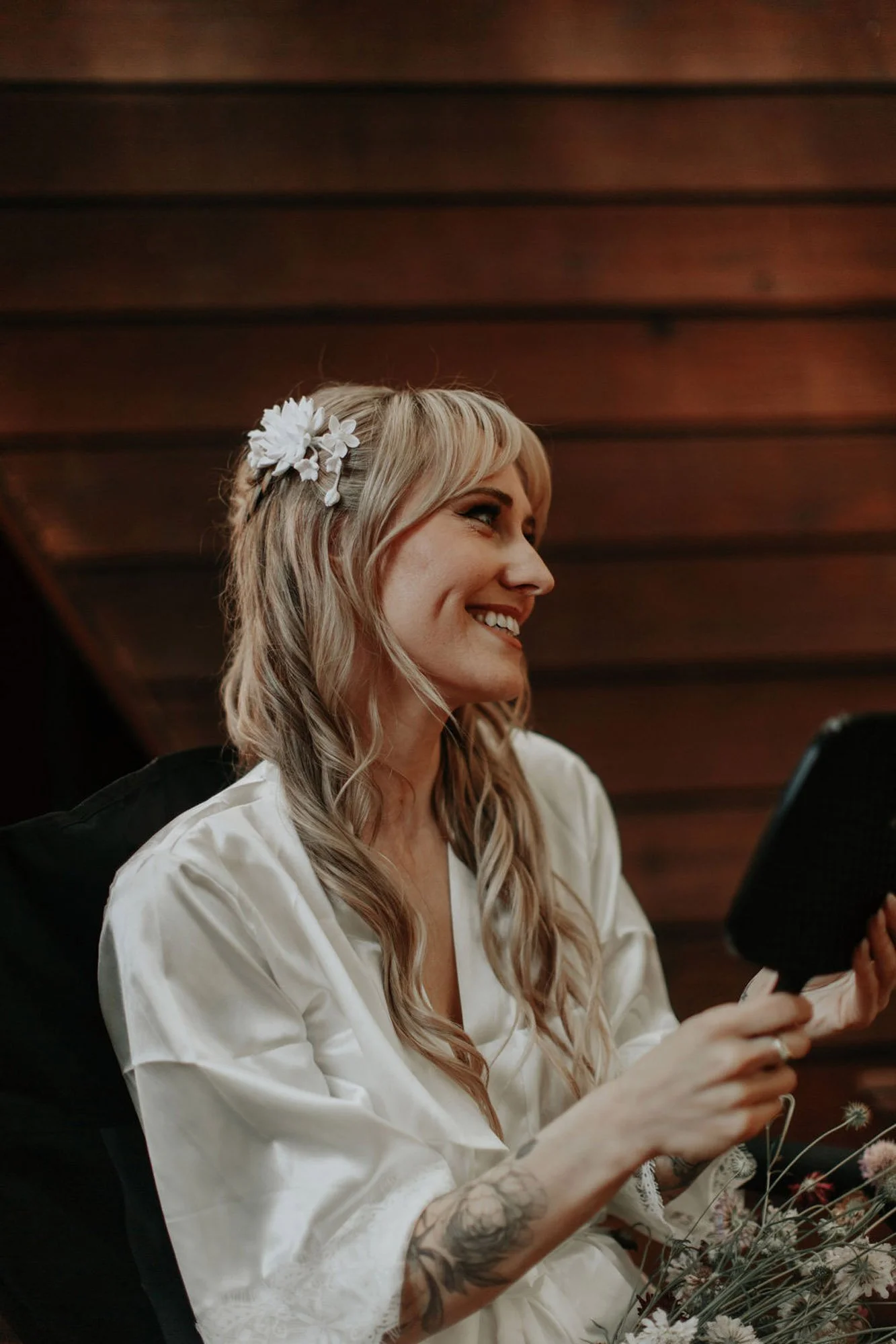 A smiling woman with blonde hair, wearing a white robe and a flower hair clip, sitting indoors in front of a wooden wall, holding a black box.