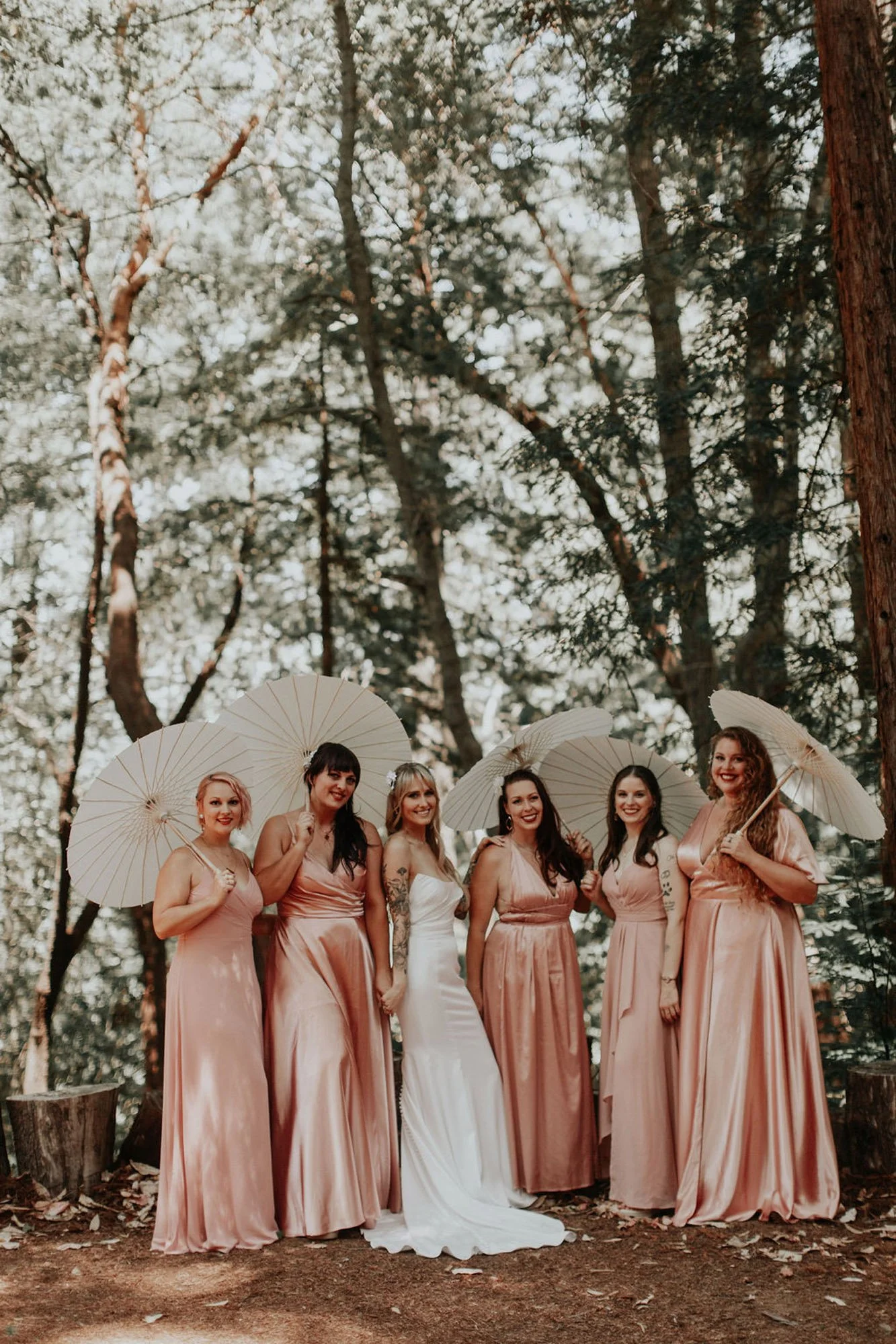A group of six women standing together in a forest, all holding white parasols and dressed in matching champagne-colored dresses, except for one woman in a white wedding gown, suggesting a bridal party.
