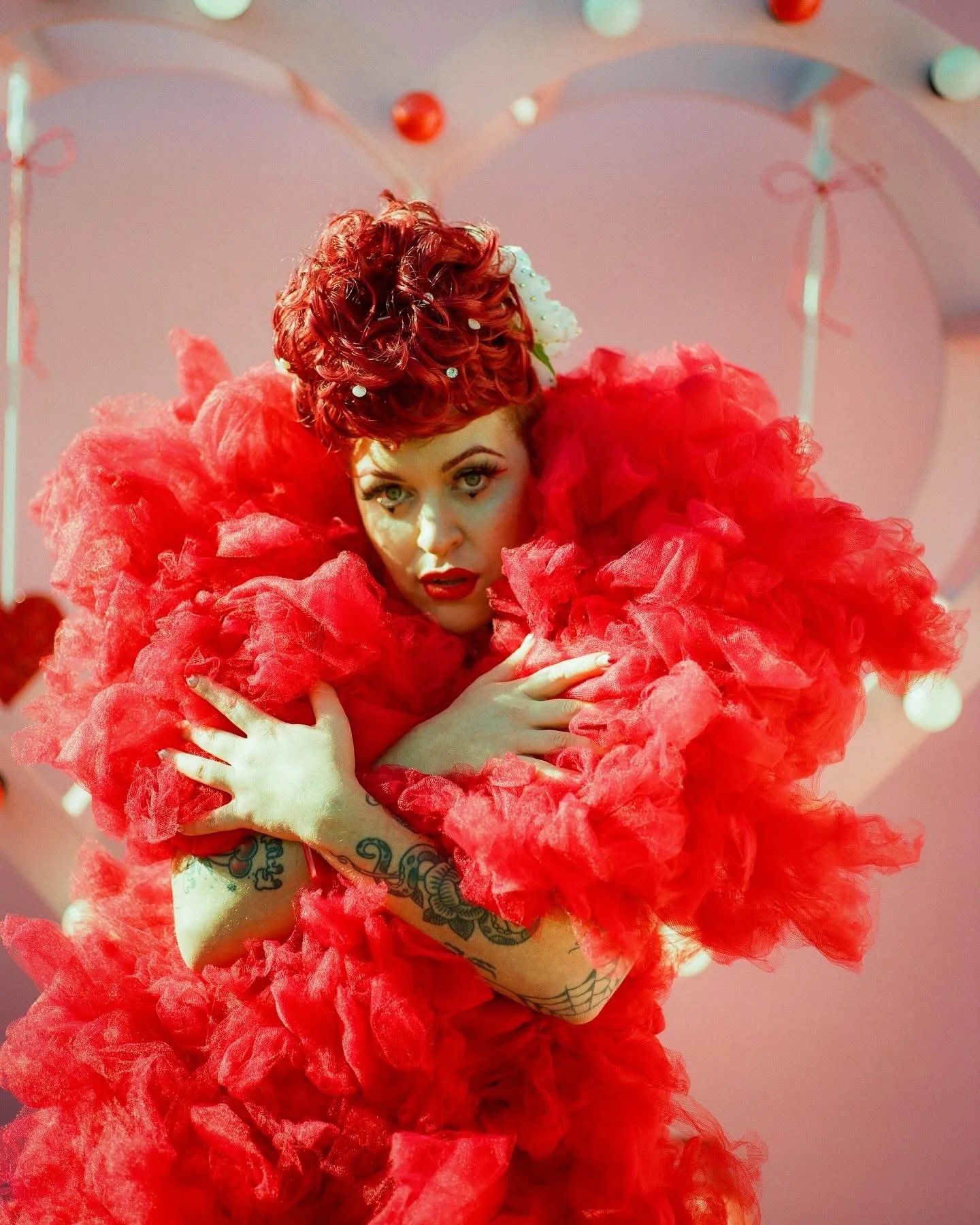 A woman with red curly hair and tattoos on her arms, wearing a red ruffled outfit, posing with her hands crossed over her chest, in front of a pink background decorated with heart-shaped balloons.