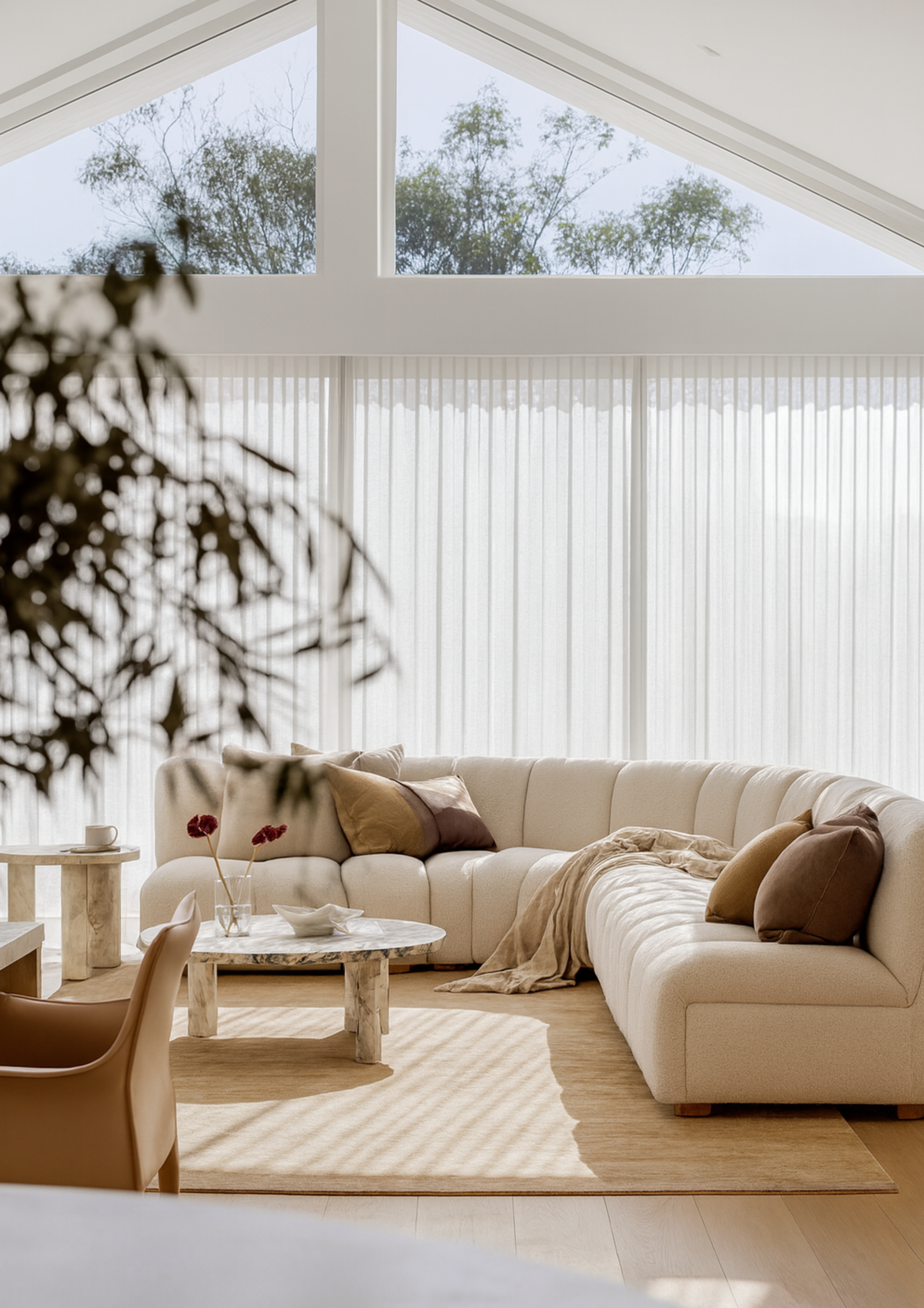 Modern living room with curved cream sofa, earthy throw pillows, marble coffee tables, floor-to-ceiling sheer curtains, and natural light, highlighting minimalist luxury interior design.