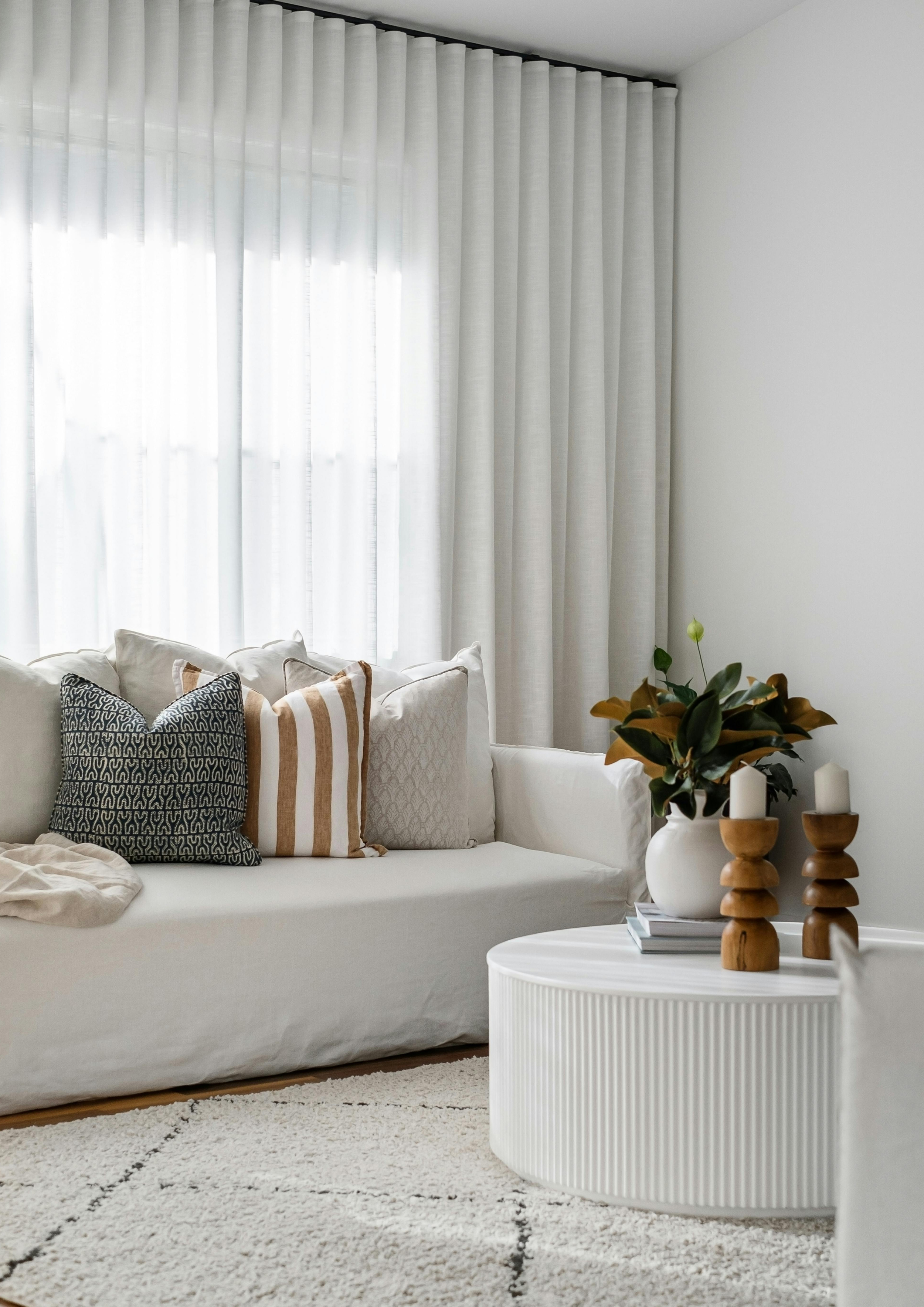 Modern living room with white sofa, patterned and striped throw pillows, round white coffee table with wooden accents, textured rug, and sheer curtains showcasing Scandinavian interior design style.