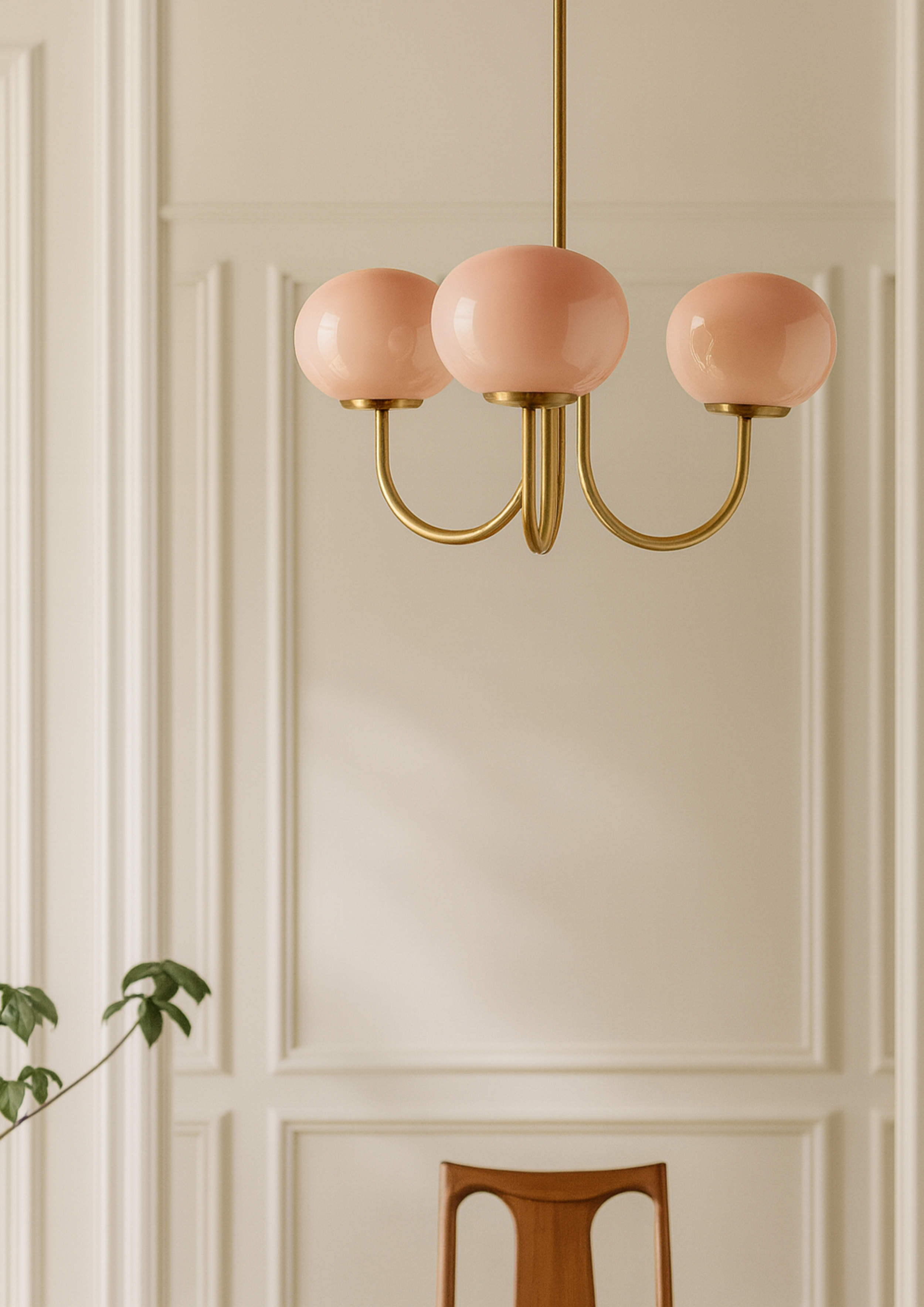 Chandelier with brass fixture and blush-pink glass globes, styled against cream paneled wall, showcasing modern Art Deco interior lighting.