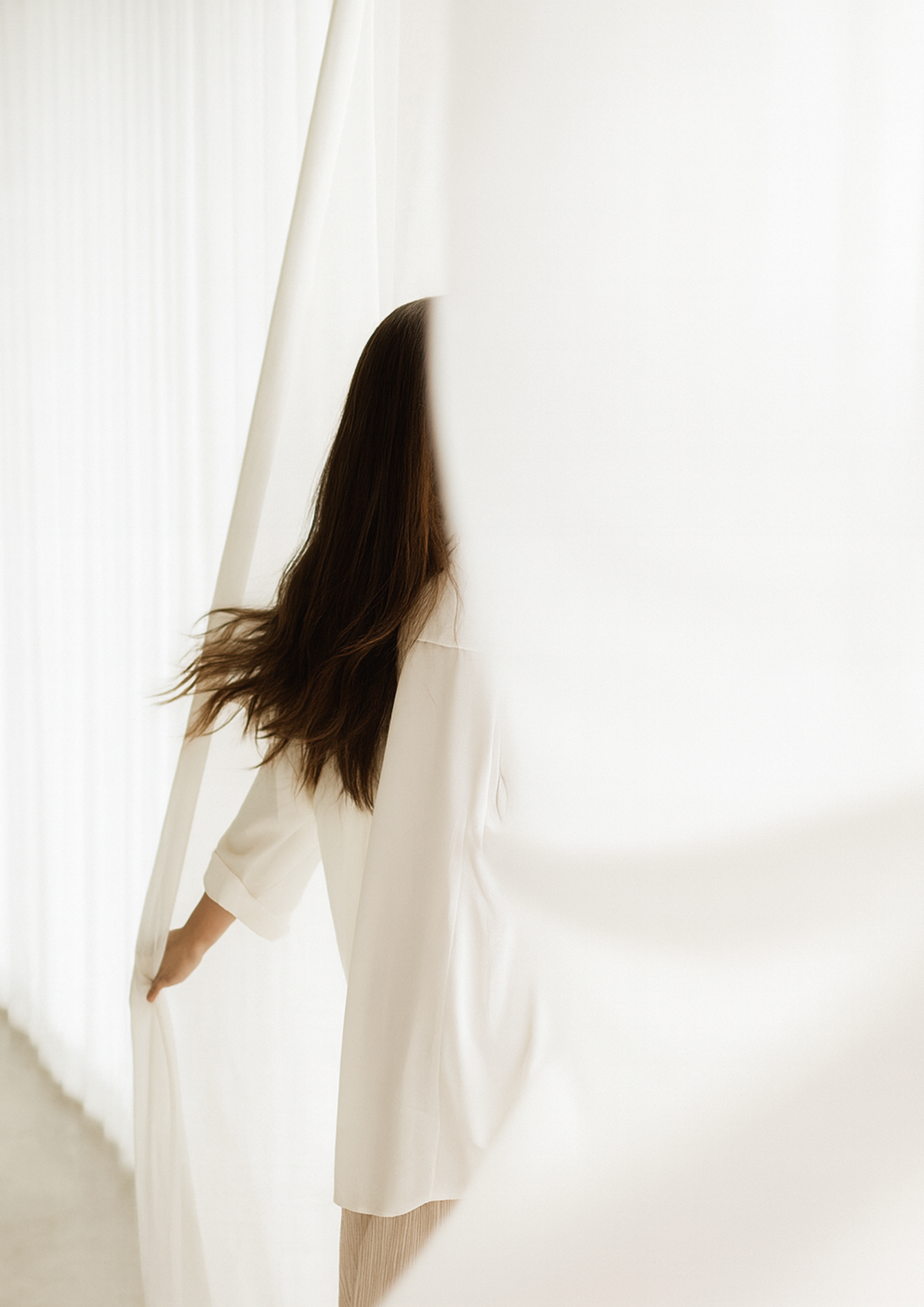 Minimalist woman with long wavy hair in a white outfit standing behind sheer curtains in a bright modern interior, symbolizing elegance, wellness, and minimalist home design.