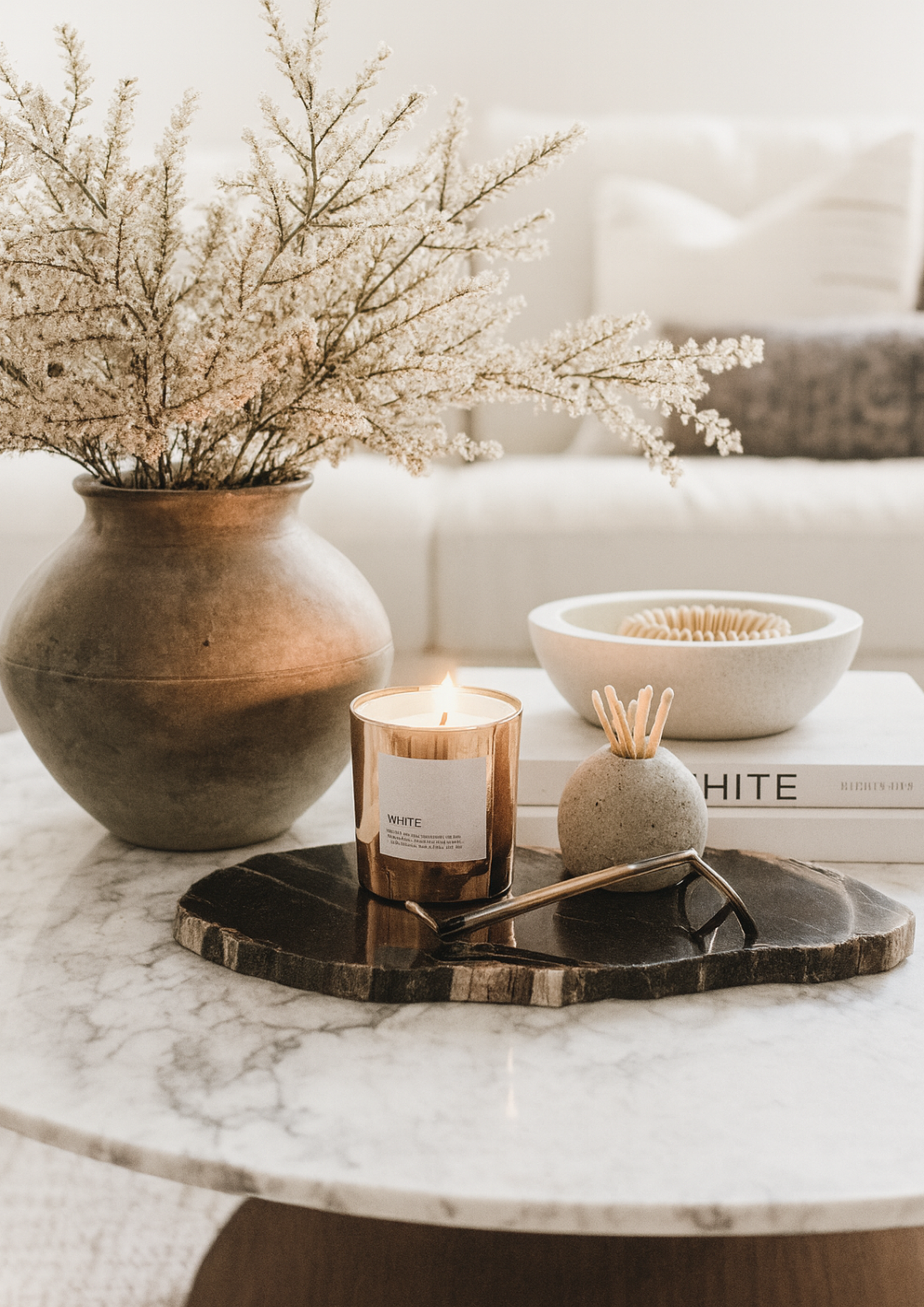 Marble coffee table styled with dried floral arrangement, lit candle, decorative accessories, and neutral-toned books, showcasing modern minimalist home decor.