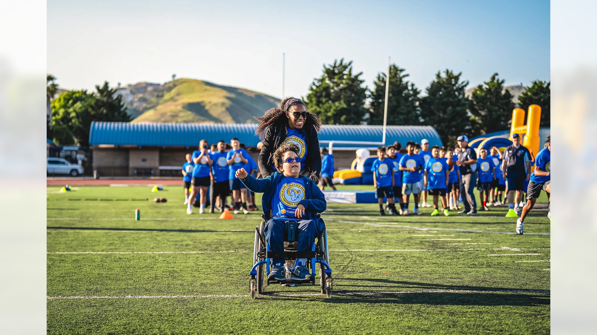 A woman pushing a young boy in a wheelchair on a football field during a youth football event, with children and adults in matching blue shirts in the background.