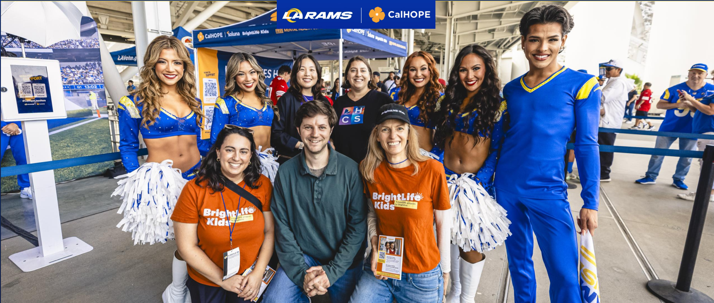 Group of people at a sports event, including cheerleaders in blue and white outfits, and casual spectators, standing together indoors with a banner above that reads 'LA Rams, CalHOPE, BrightLife Kids'.