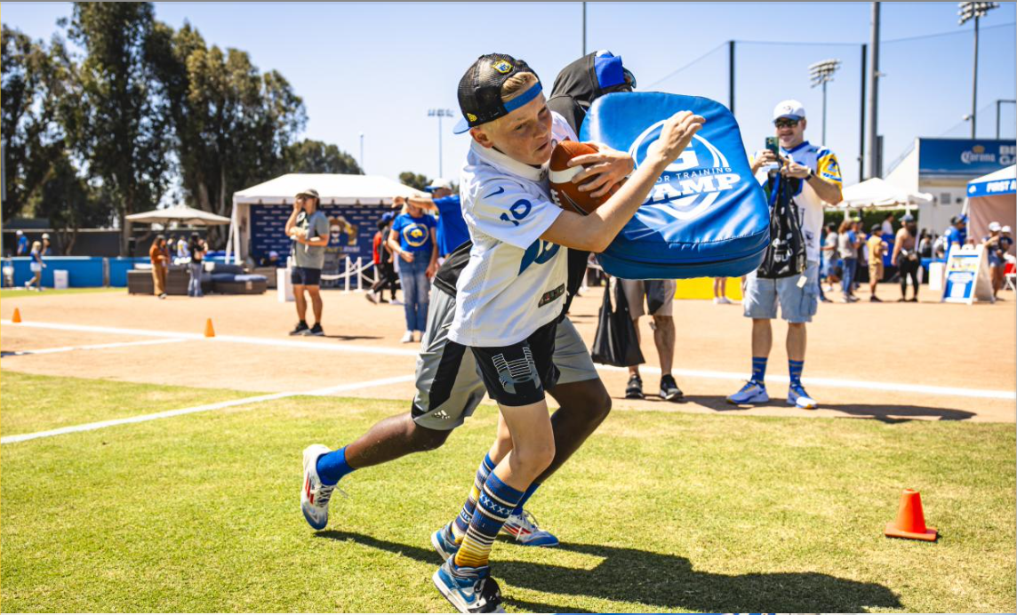 A young boy wearing a baseball cap and patterned socks runs with a football while carrying a blue training bag on a sports field during daytime, with other people and tents in the background.