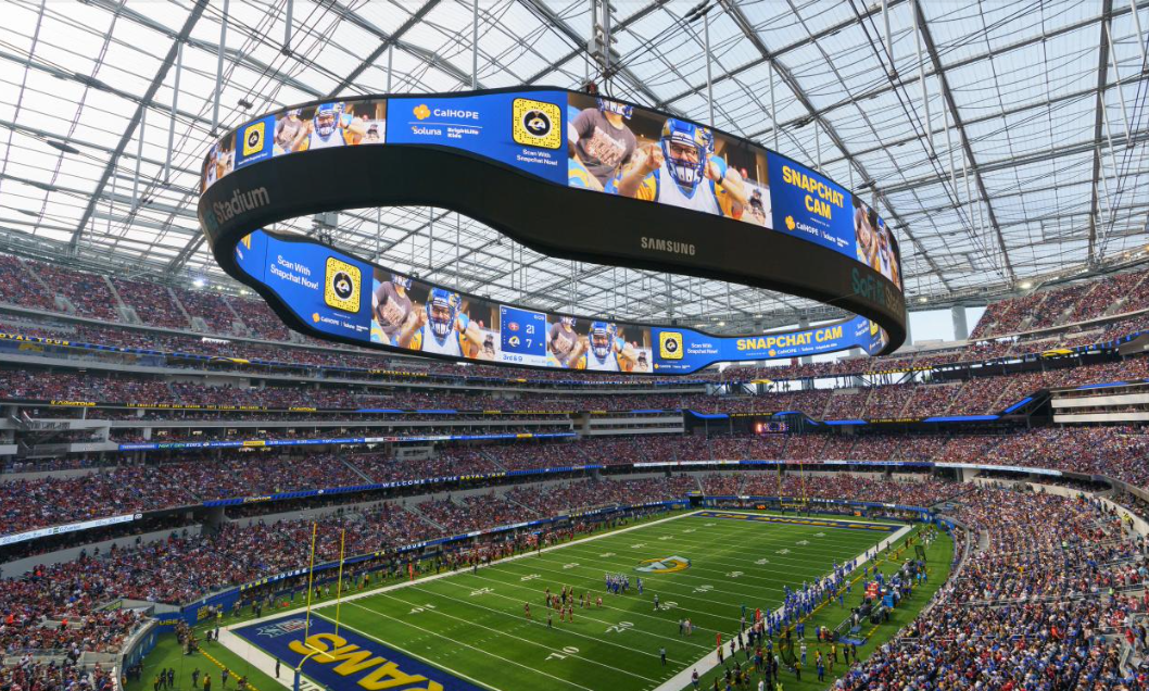 Inside a large modern football stadium filled with spectators, with a view of the field and a large digital scoreboard overhead. The scoreboard displays a promotional ad for Snapchat CAM and shows a football game in progress.