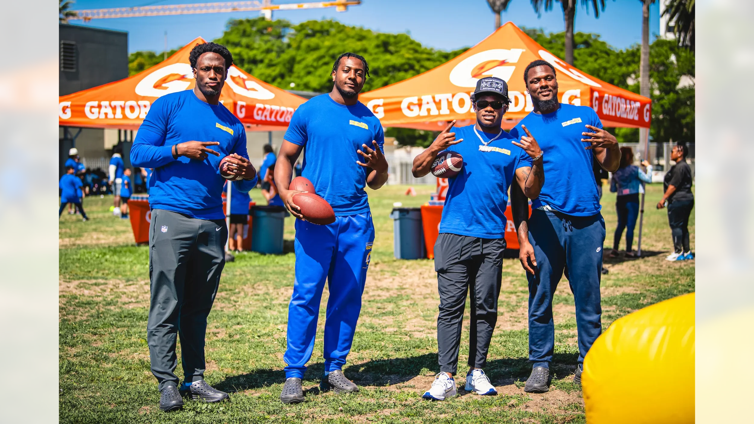 Four men wearing blue shirts and dark pants standing on grass at outdoor Gatorade event, holding footballs, with Gatorade tents and people in the background.