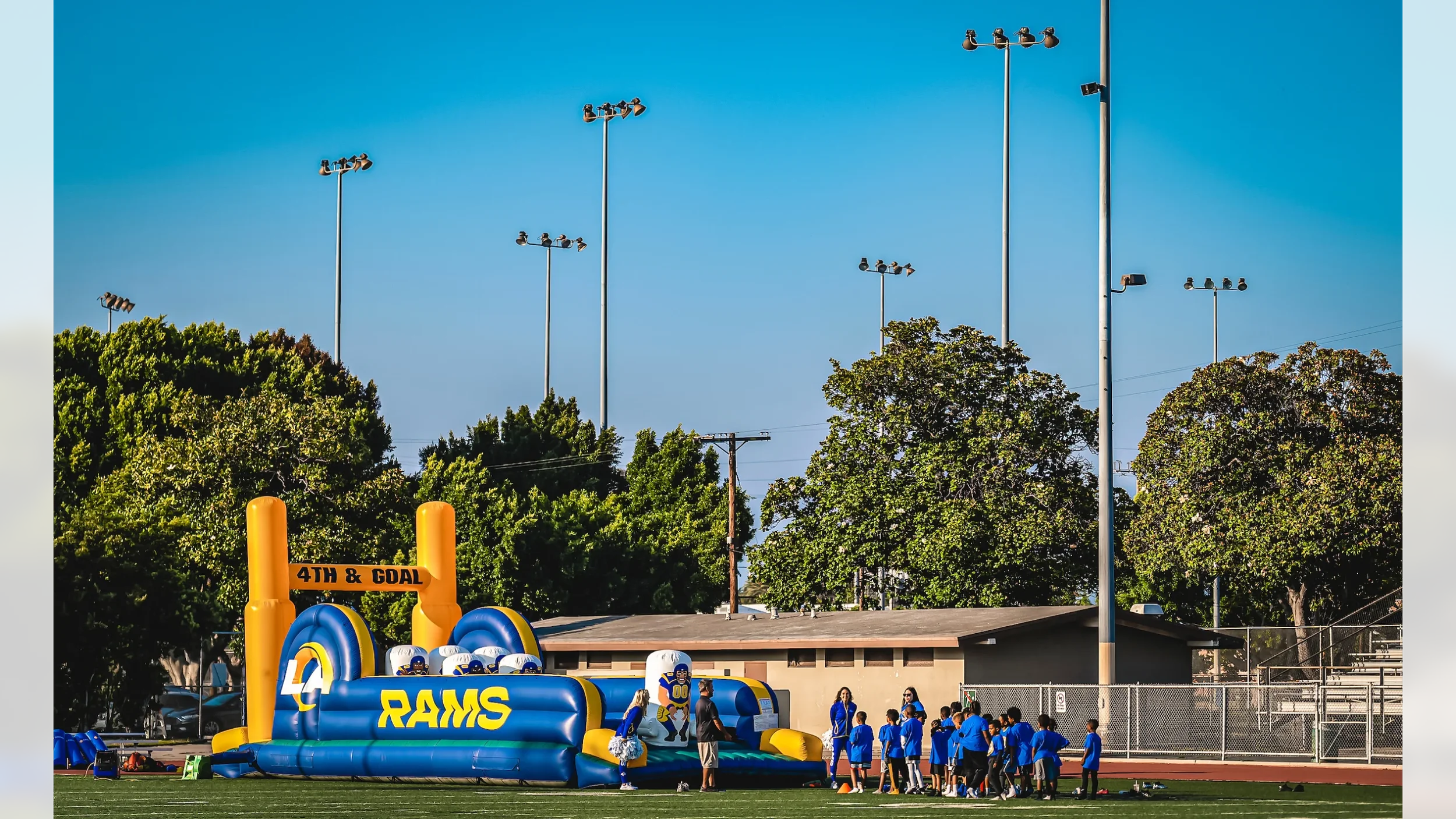 Inflatable football-themed obstacle course on a school field with children in blue shirts, coaches, and coaches, with trees and light poles in the background.