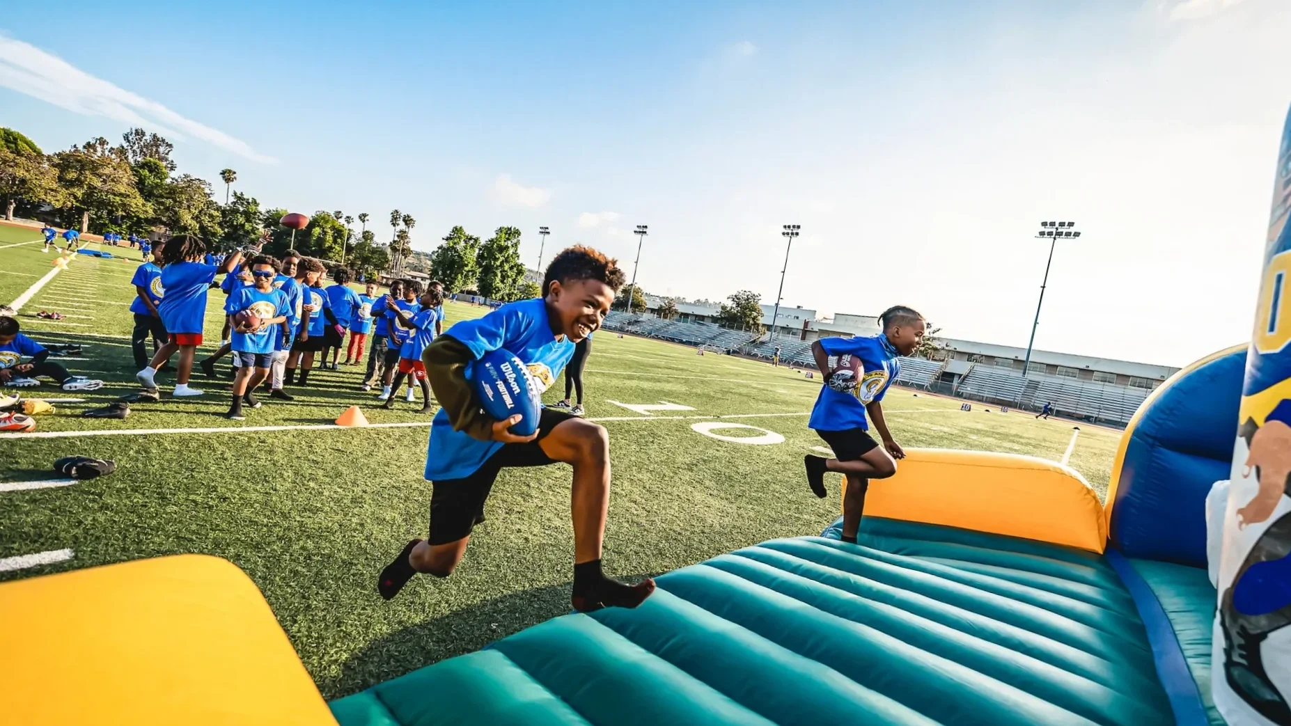 Children participating in a sports activity on a football field, with some jumping over an inflatable hurdle, wearing blue shirts.