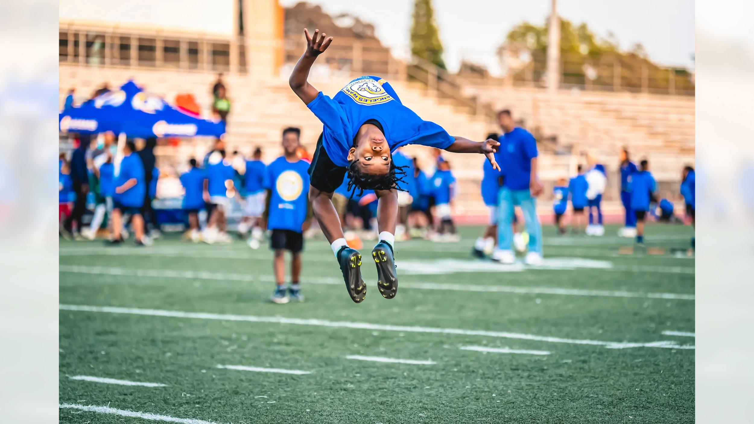 A young boy in a blue sports uniform is mid-air performing a flip on a football field during a sporting event with several other children and adults in similar uniforms in the background.