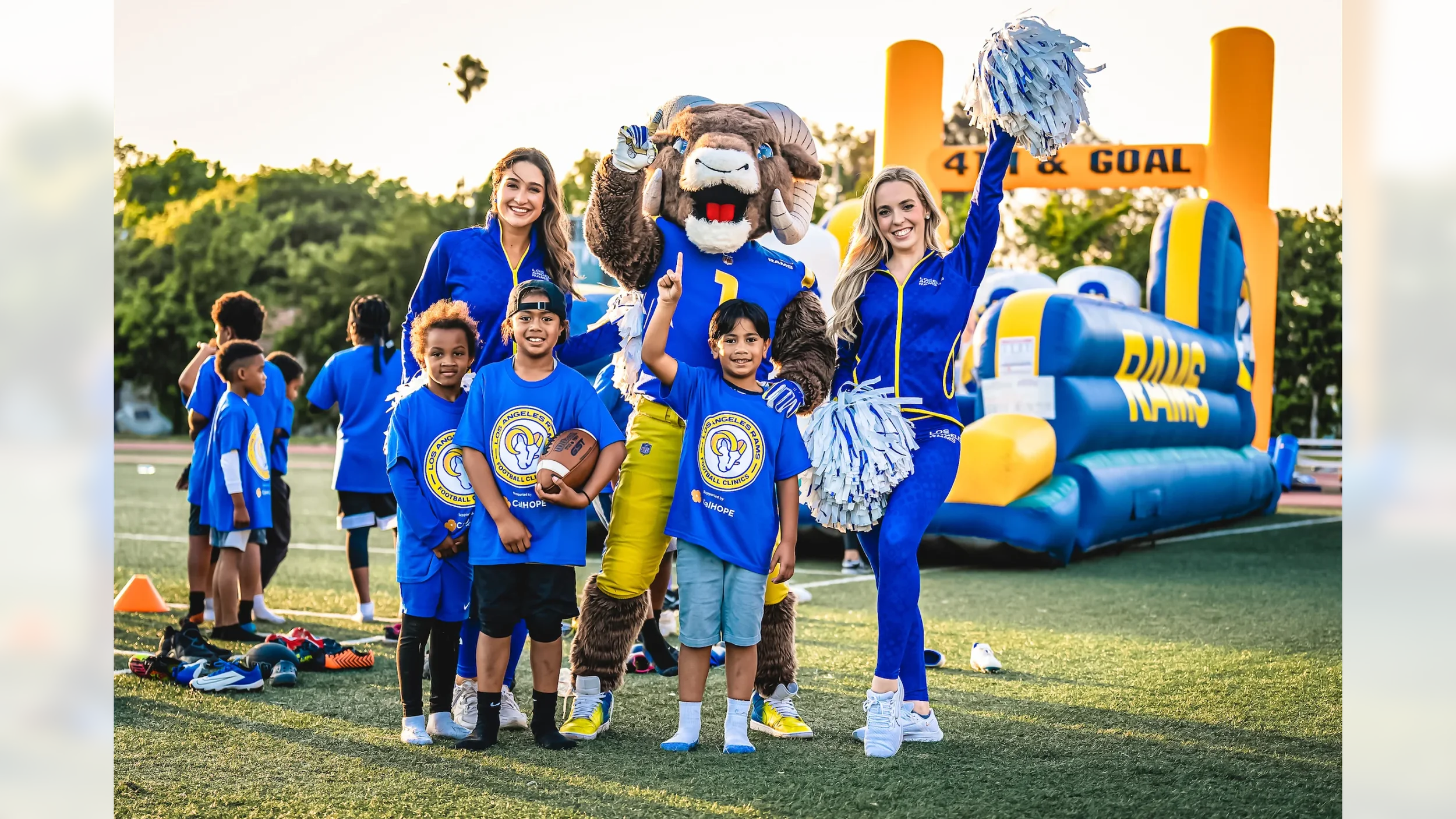 A group of young children and cheerleaders pose with a lion mascot on a football field during a community event. The children are wearing blue Los Angeles Rams shirts, and the cheerleaders are in blue uniforms with pom-poms. An inflatable Rams footba