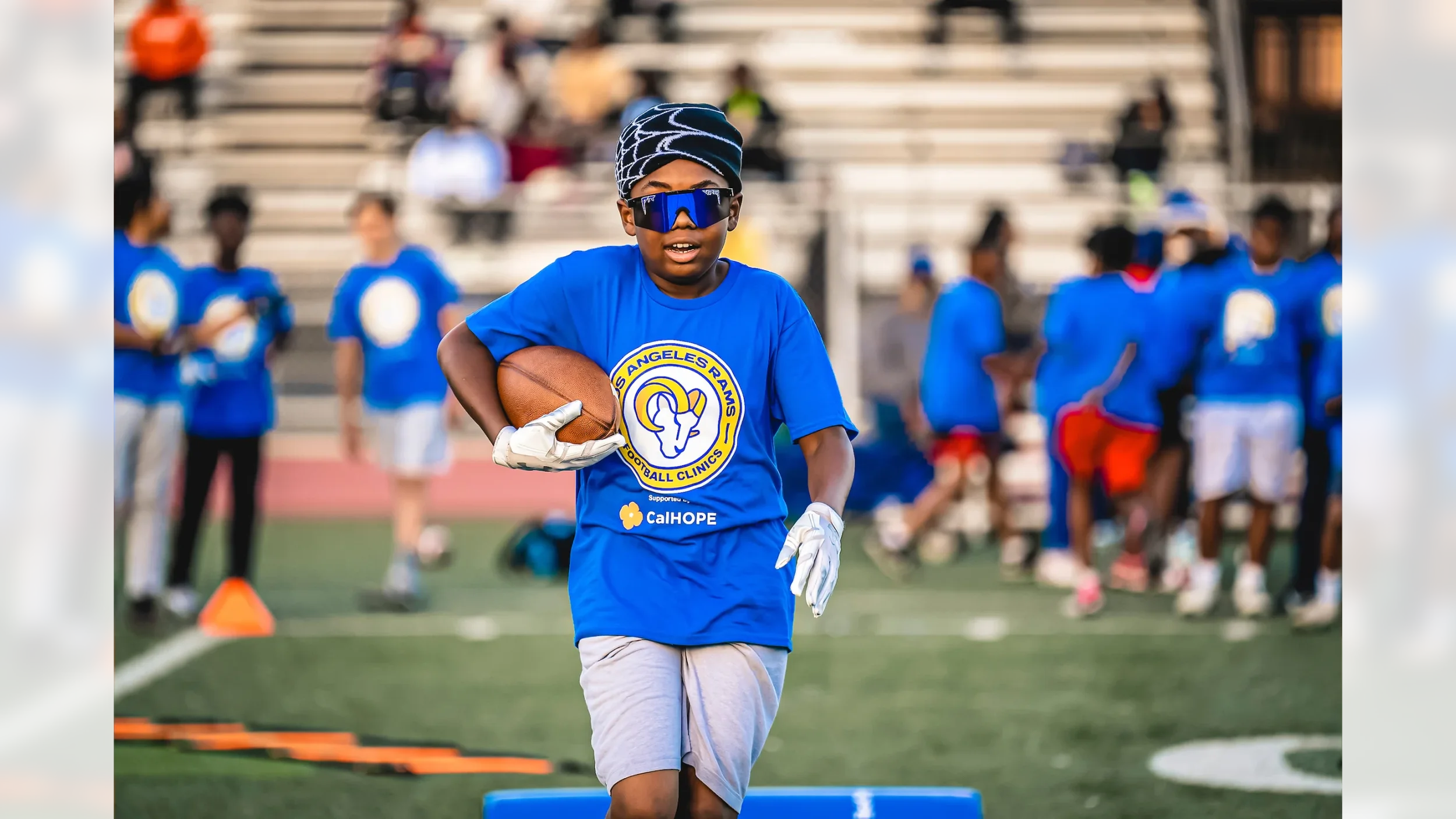 A young boy in a blue Los Angeles Rams football clinic shirt, sunglasses, and head covering running on a football field while holding a football, with other children and people in the background.