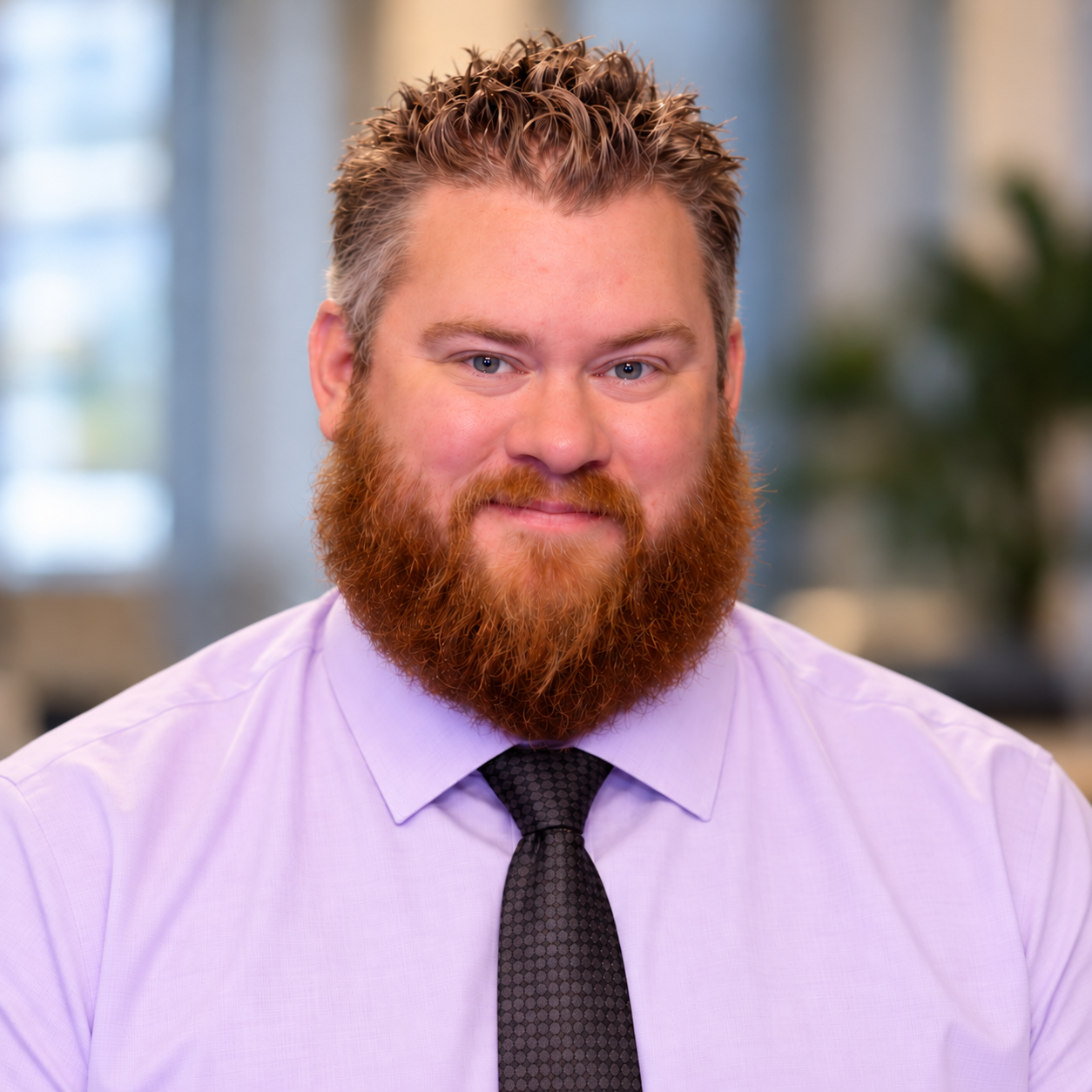 A man with a beard and mustache, wearing a checkered shirt and a dark tie, smiling against a white background.