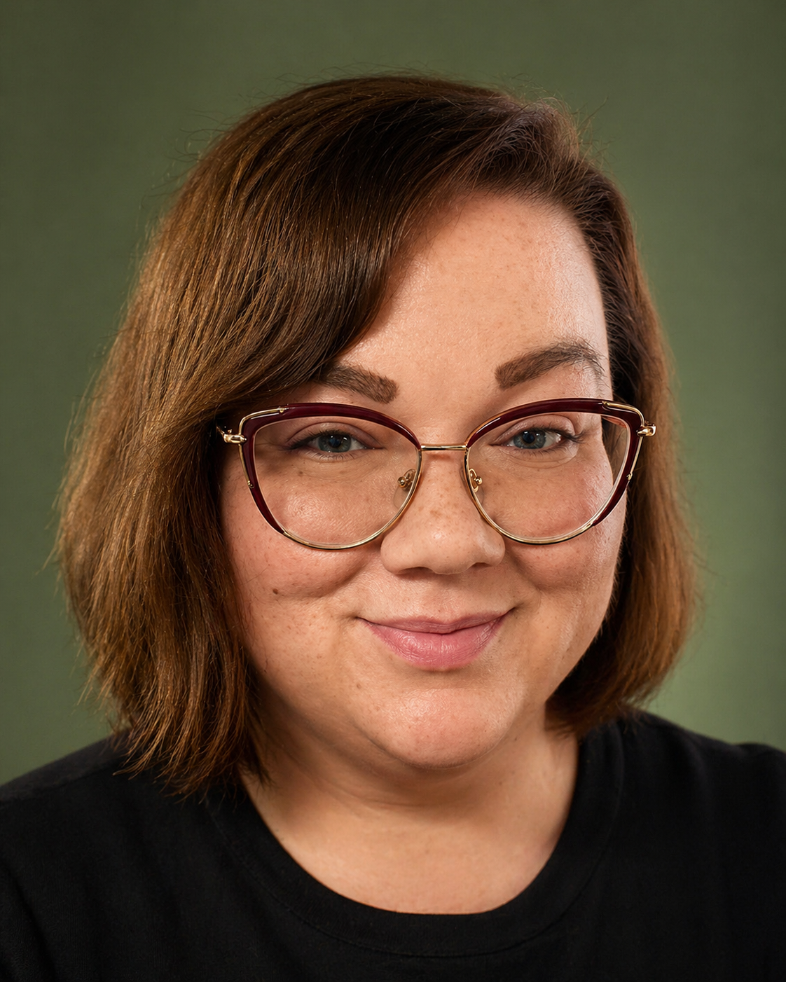A woman with shoulder-length brown hair, wearing large pink glasses and a black T-shirt featuring celestial and mystical designs, smiling at the camera.