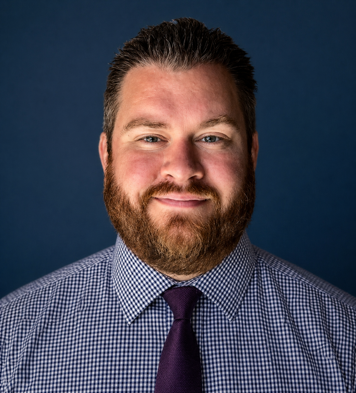 A man with a beard and mustache, wearing a checkered shirt and a dark tie, smiling against a white background.