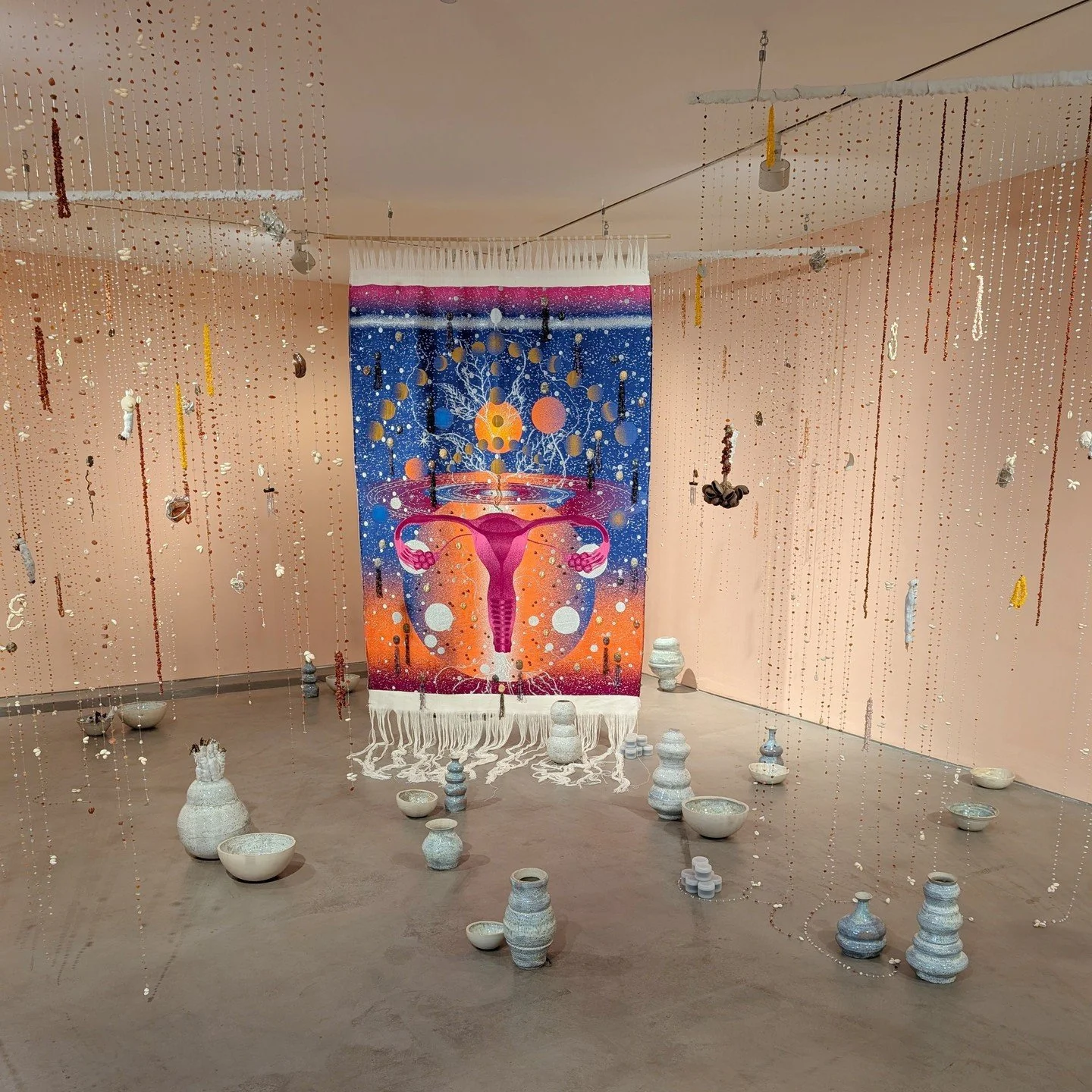 A cosmic textile artwork with a uterus at its centre, ceramic vessels on the floor and lines of beads hanging from the ceiling. Adapting traditions from her Creole heritage and reflecting on stories of intergenerational trauma she has created immersi