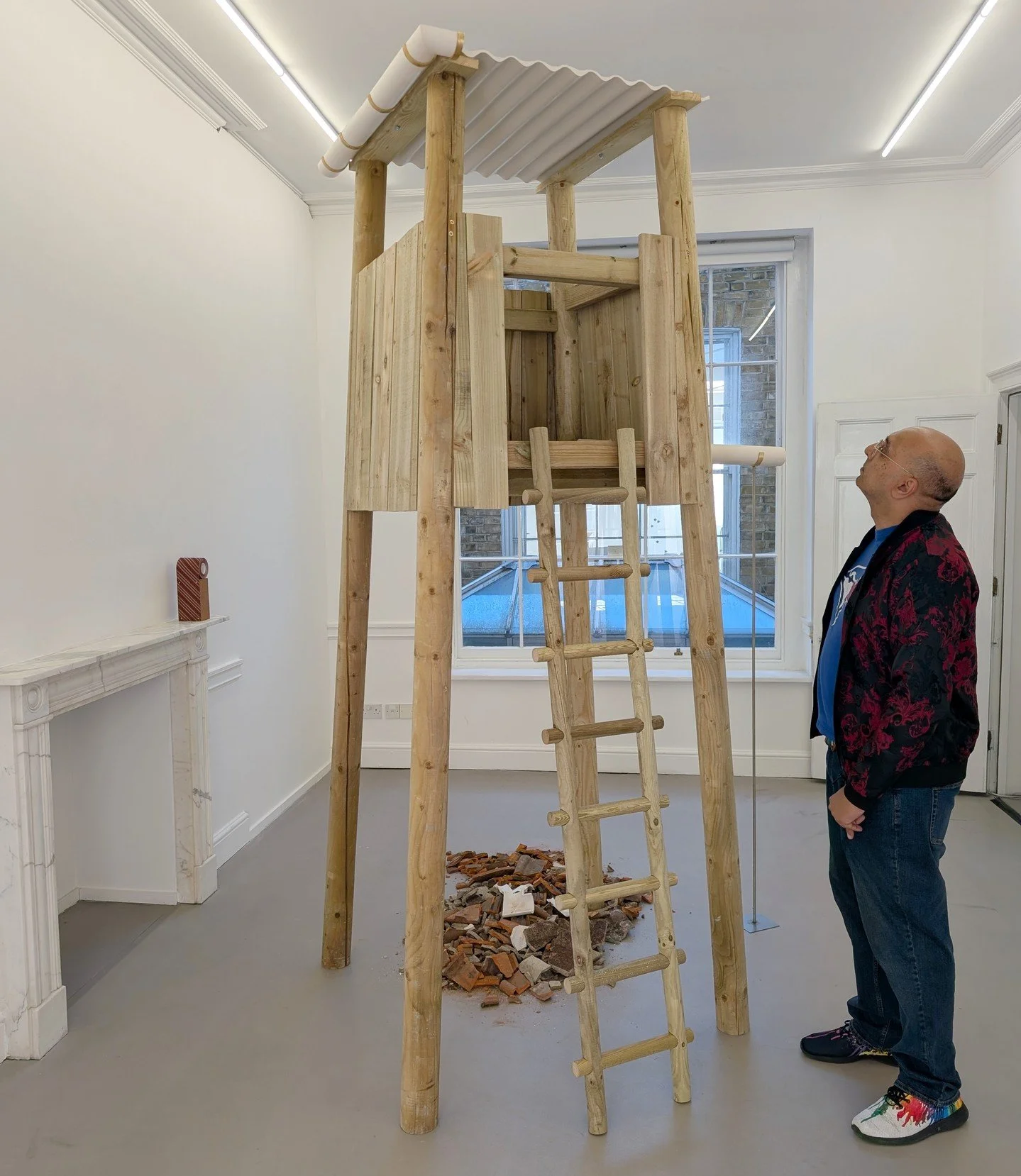 Two birdwatching towers made from reclaimed wood dominate the space, broken tiles on the floor suggest the tops have fallen off, only to be replaced with a Jesmonite roof. Around them are abstract works on sandpaper belts made purely by applying diff