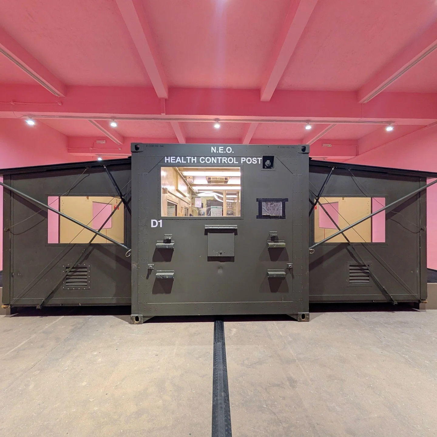 Enter the space, and a militaristic structure dominates. We can walk inside, and despite its oppressive look, it&rsquo;s been used as a mobile health unit. Surrounding it, the walls are painted a particular shade of pink designed to soothe people and