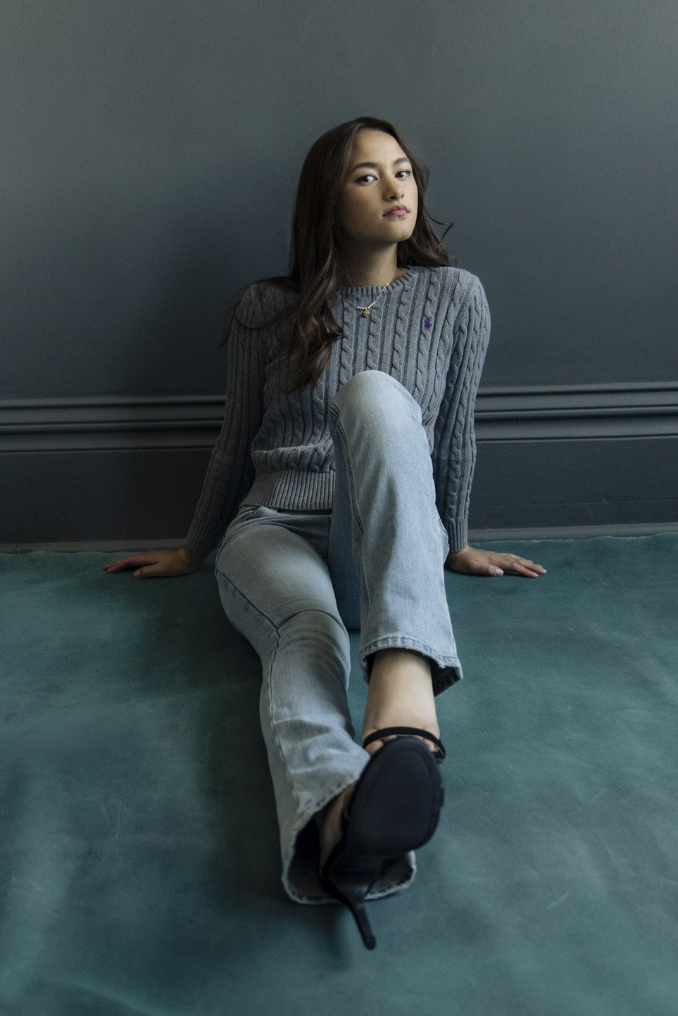 Full-length modeling portfolio portrait of Gretchen in a gray sweater seated against a dark teal studio wall.