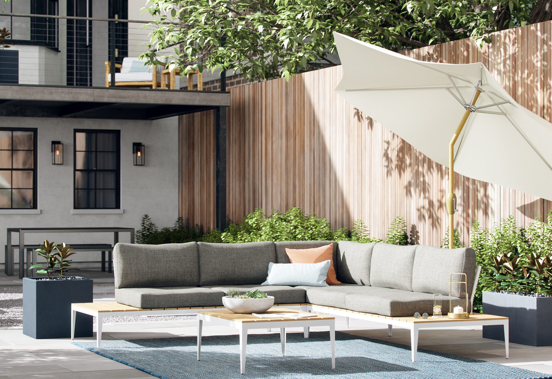 Outdoor patio area with a large L-shaped gray sofa, a white coffee table with a bowl and decorative items, potted plants, a large white umbrella, and a wooden fence with green shrubbery in the background.