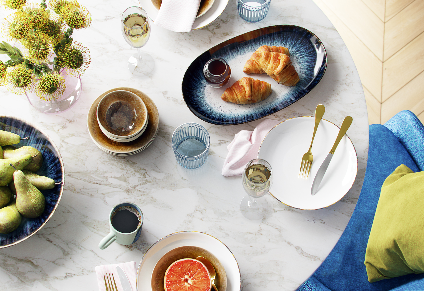 Overhead view of a breakfast table with croissants, a grapefruit, coffee, juice, and water, set with plates, cutlery, and flowers.