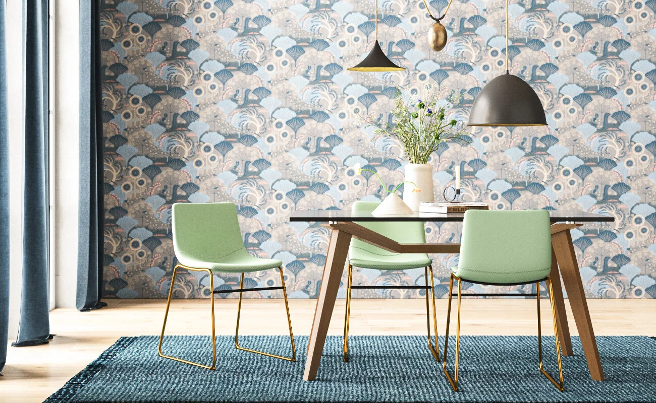 Modern dining room with wooden table, three light green chairs with gold legs, a blue patterned rug, floral wallpaper, black pendant lights, a white vase with flowers, a candle, and books.