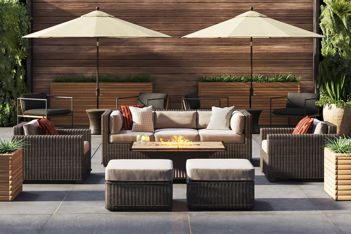 Outdoor patio with wicker furniture, beige sofa with pillows, two armchairs, a fire pit, two umbrellas, and potted plants against a wood fence.