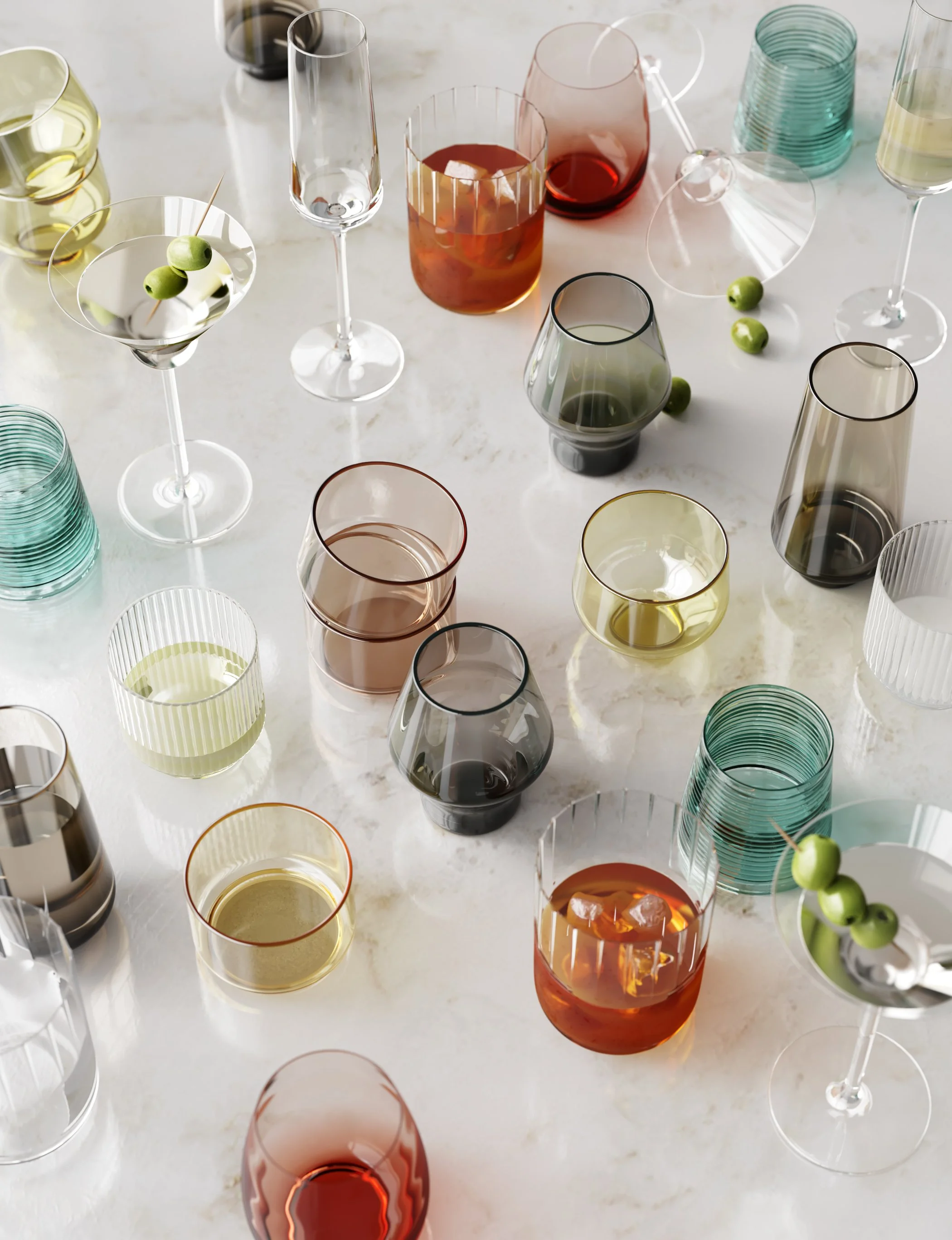 A variety of colorful glasses, some empty and some filled with liquids, are arranged on a white marble surface. Some glasses have olives and skewers inside, while others are stacked or positioned separately.